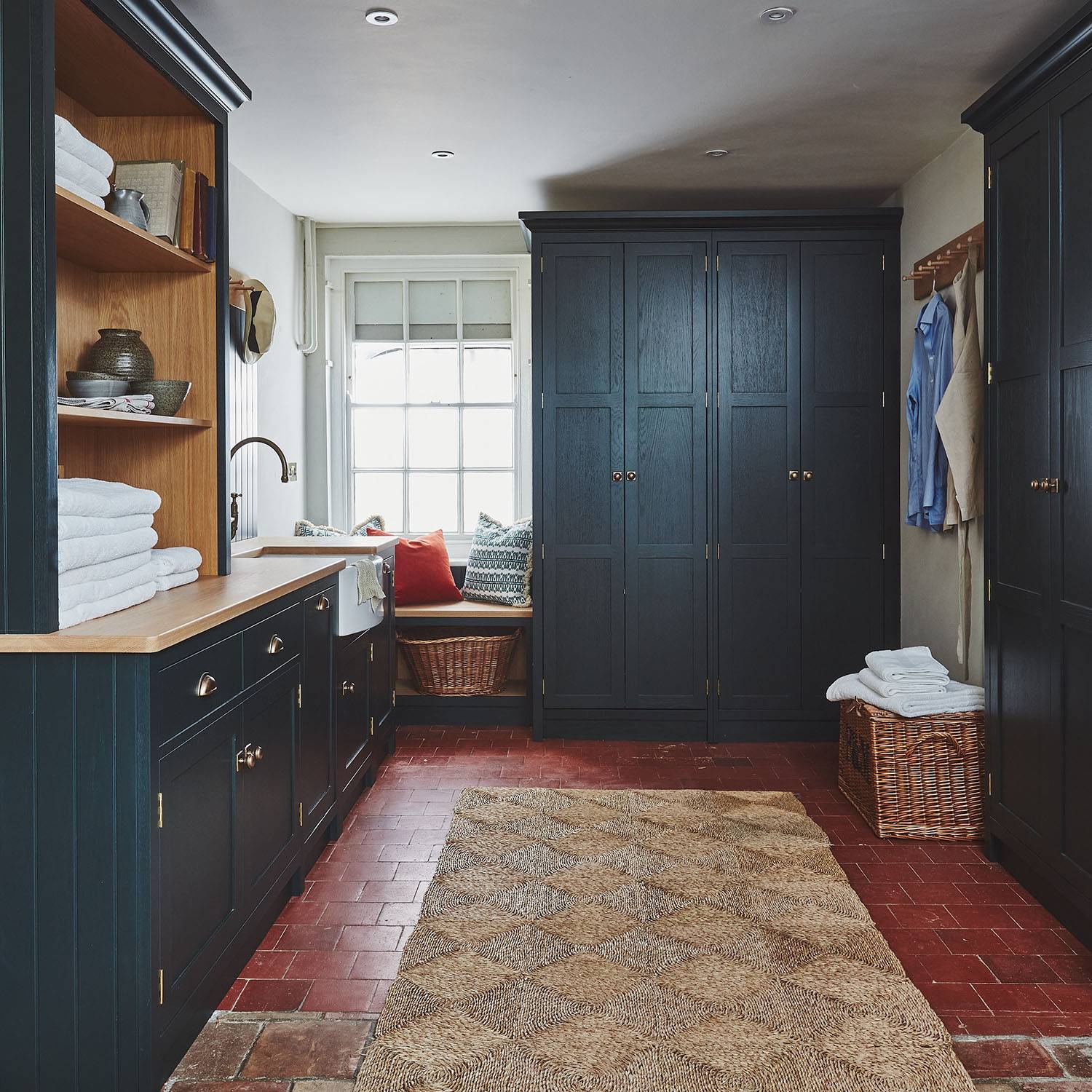 A cozy laundry room with dark cabinets, a countertop with folded towels, a stacked washer and dryer, a window seat with cushions, and a woven rug on a red tile floor. Shirts hang on pegs along the wall.