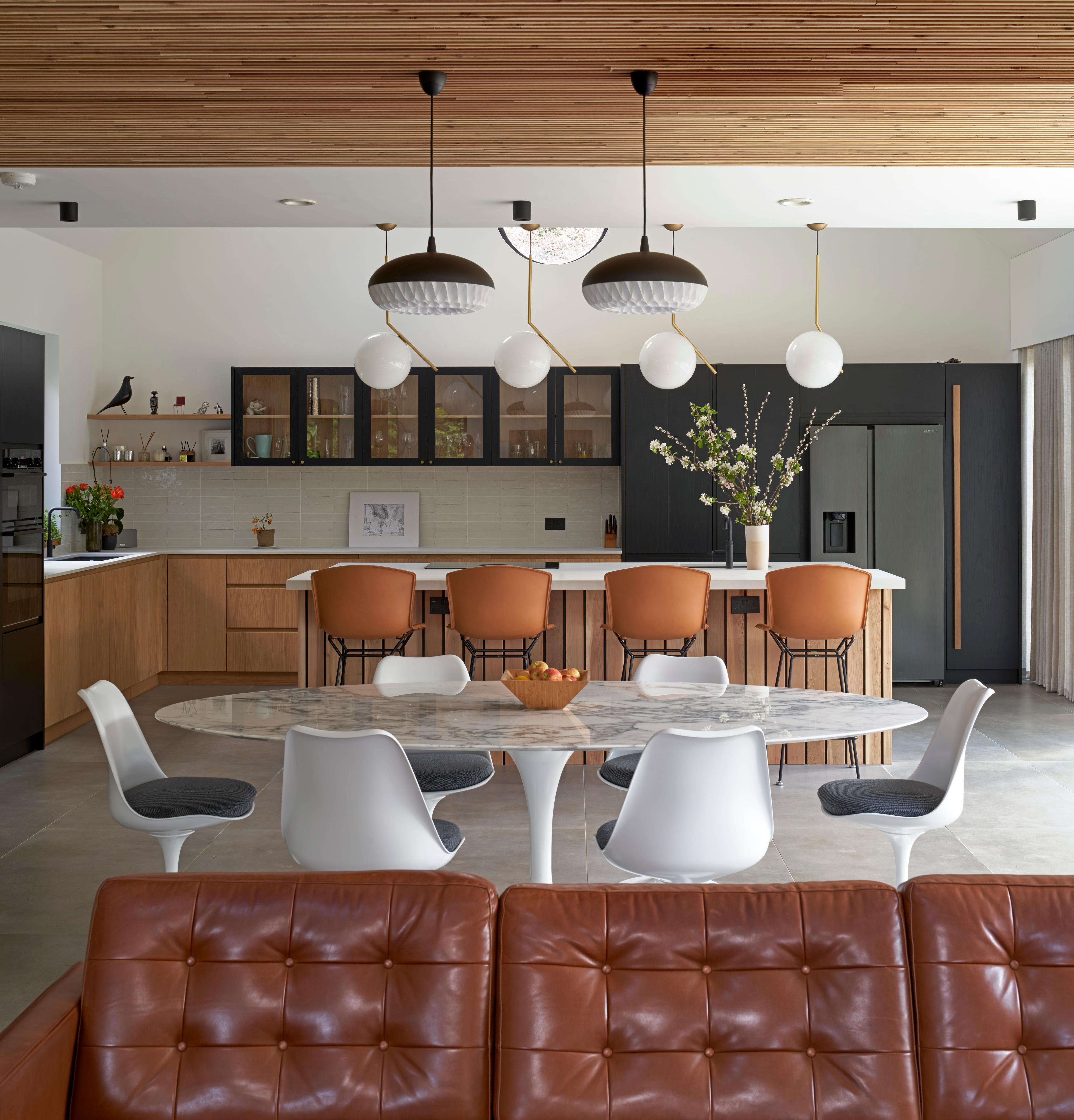 Modern kitchen with natural wood cabinetry, a large island with an induction cooktop, and light gray countertops. A window with plants on the sill allows natural light to brighten the space. Black appliances, pendant lights, and minimalistic decor complete the sleek look.