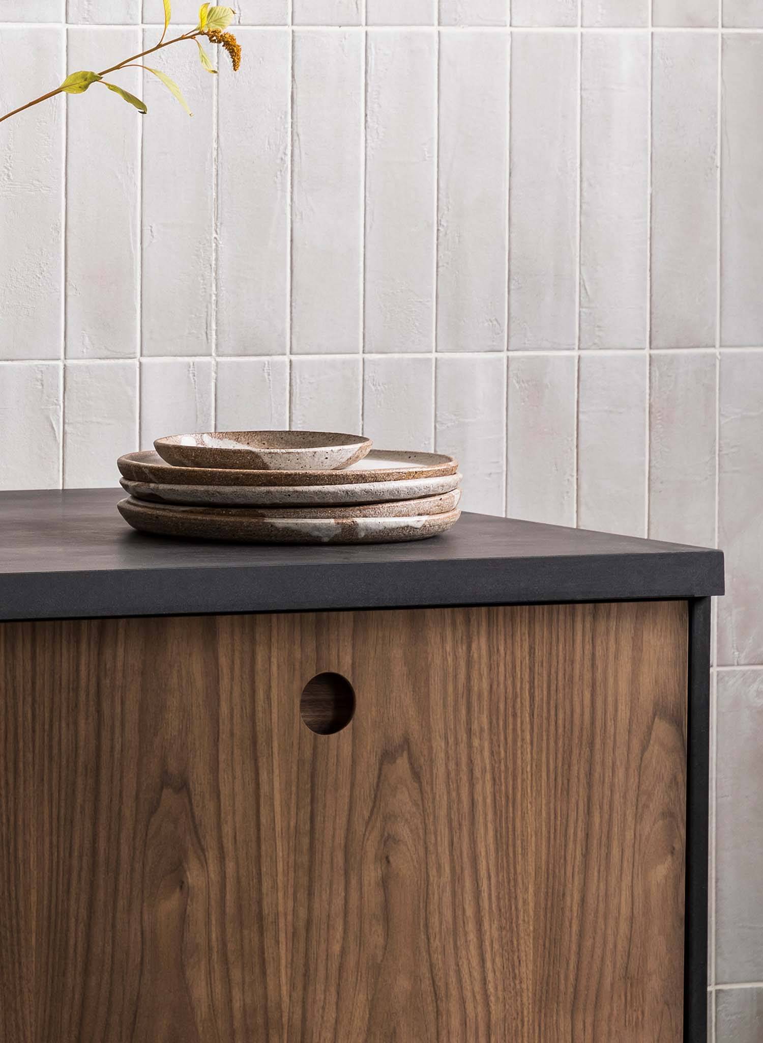 A stack of ceramic plates sits on a dark countertop above a wooden cabinet with a round hole as a handle, against a light tiled wall with vertical lines. A branch with yellow-green leaves hangs in the top left corner.
