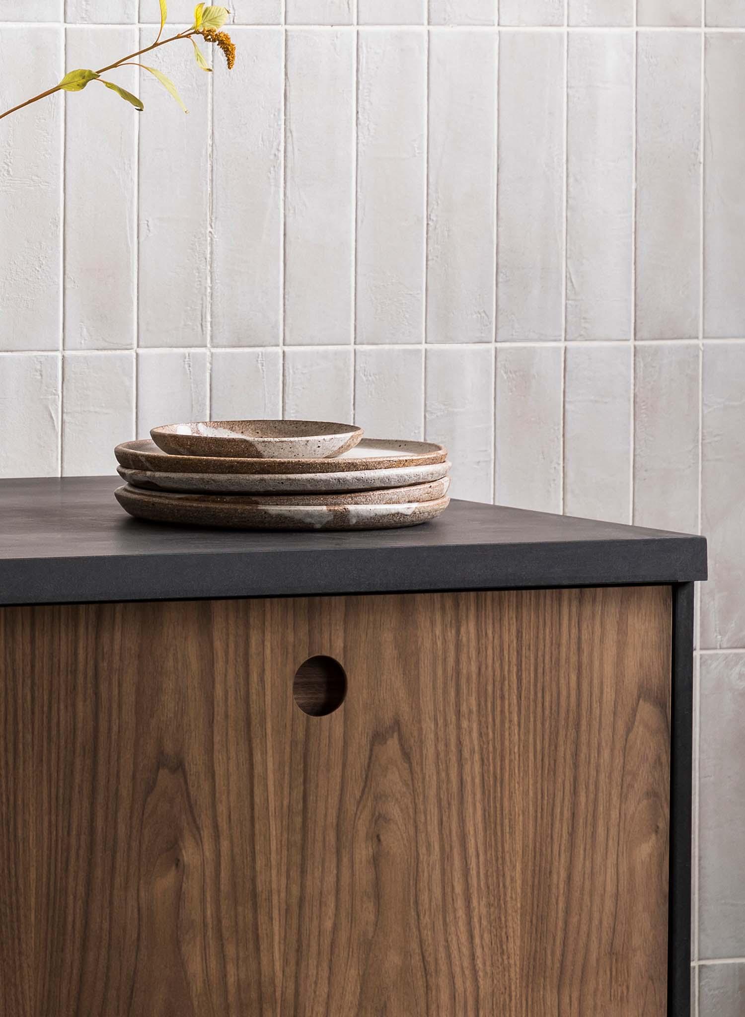 A stack of ceramic plates sits on a dark countertop above a wooden cabinet with a round hole as a handle, against a light tiled wall with vertical lines. A branch with yellow-green leaves hangs in the top left corner.