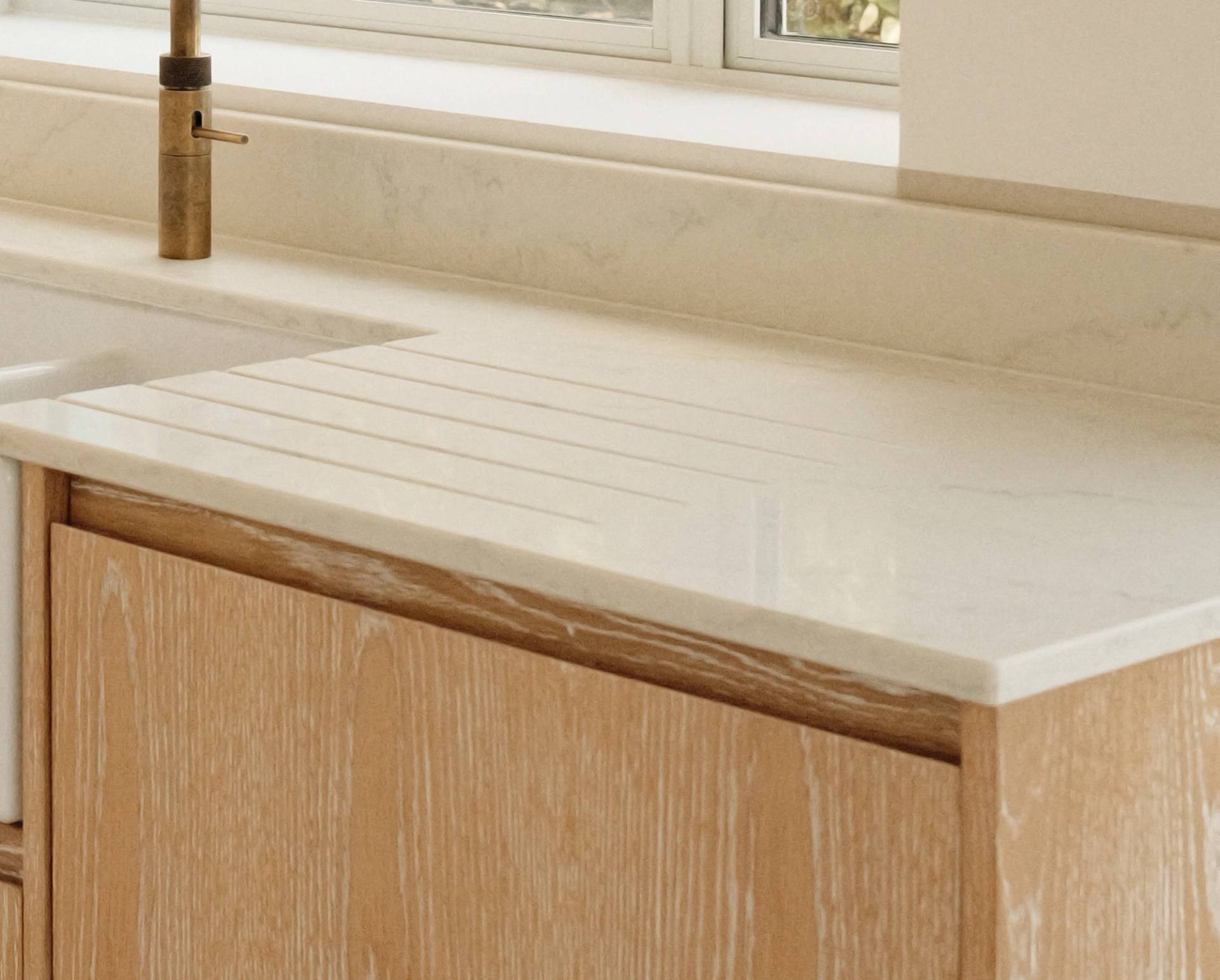 Light wood kitchen cabinet with a white marble countertop, featuring an integrated draining board with grooves next to a sink and a brass faucet, set against a light-colored backsplash under a window.