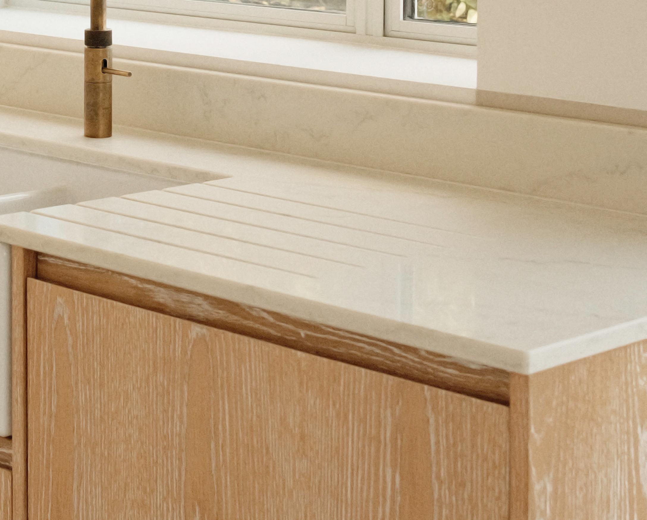 Light wood kitchen cabinet with a white marble countertop, featuring an integrated draining board with grooves next to a sink and a brass faucet, set against a light-colored backsplash under a window.