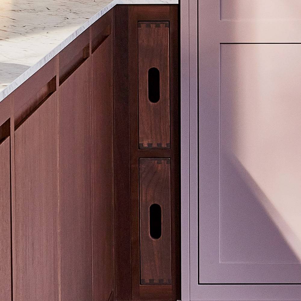 Close-up of two wooden pull-out drawers with oval cut-out handles, set into the side of a kitchen island next to a panel with a light purple finish and a marble countertop above.