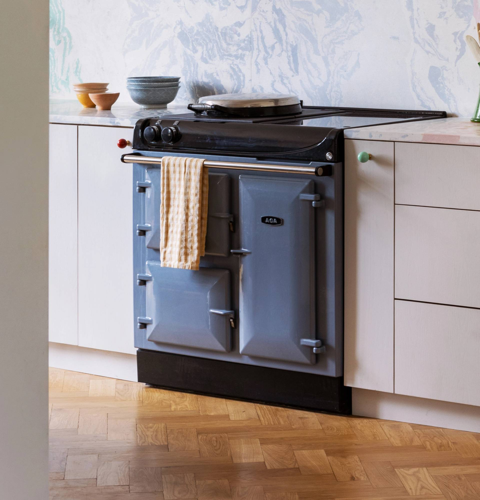 A blue AGA stove with a striped towel hanging from the handle is set against a white kitchen cabinet. There are bowls and a vase on the countertop. The floor has a wooden parquet pattern.