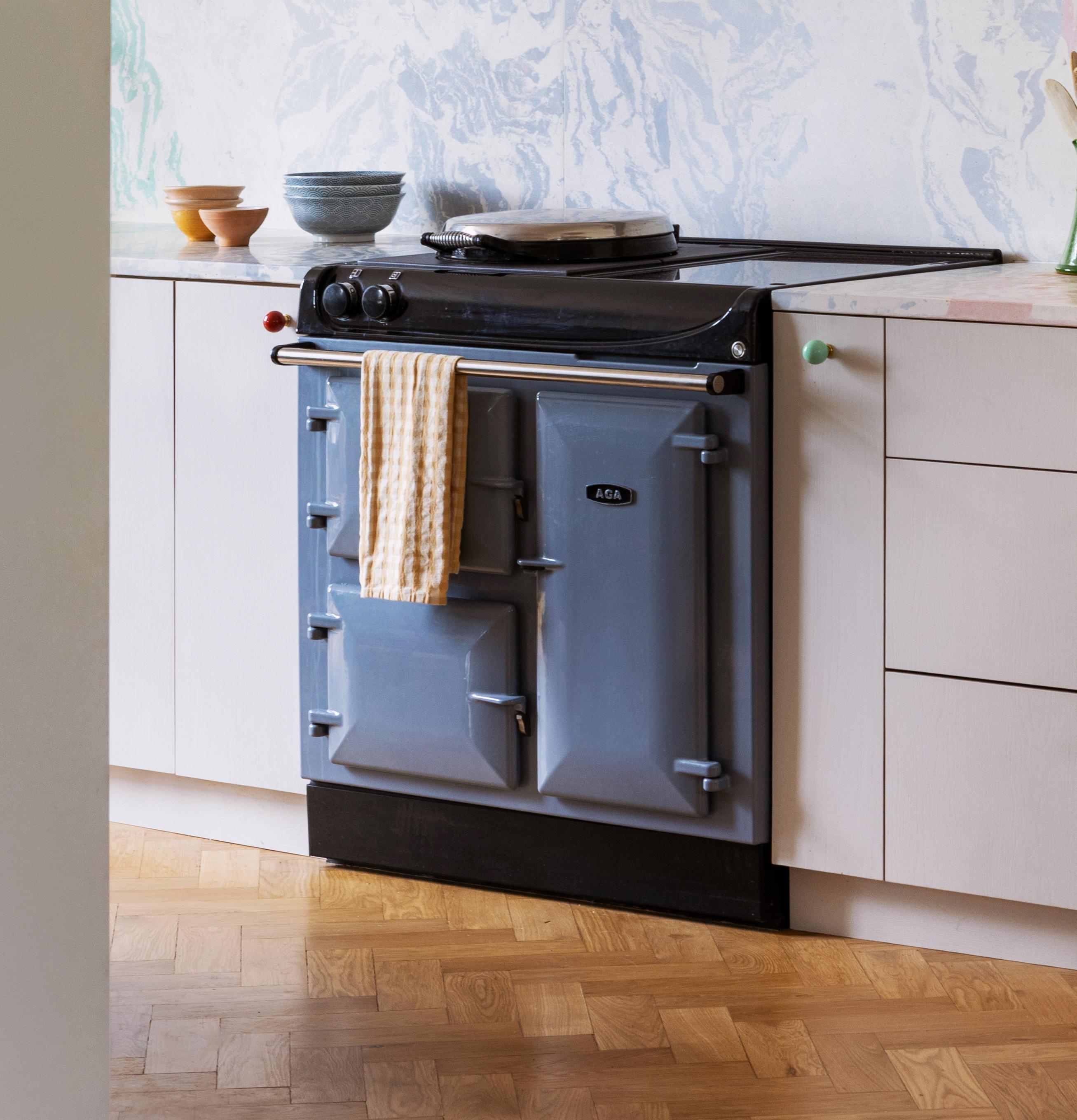 A blue AGA stove with a striped towel hanging from the handle is set against a white kitchen cabinet. There are bowls and a vase on the countertop. The floor has a wooden parquet pattern.