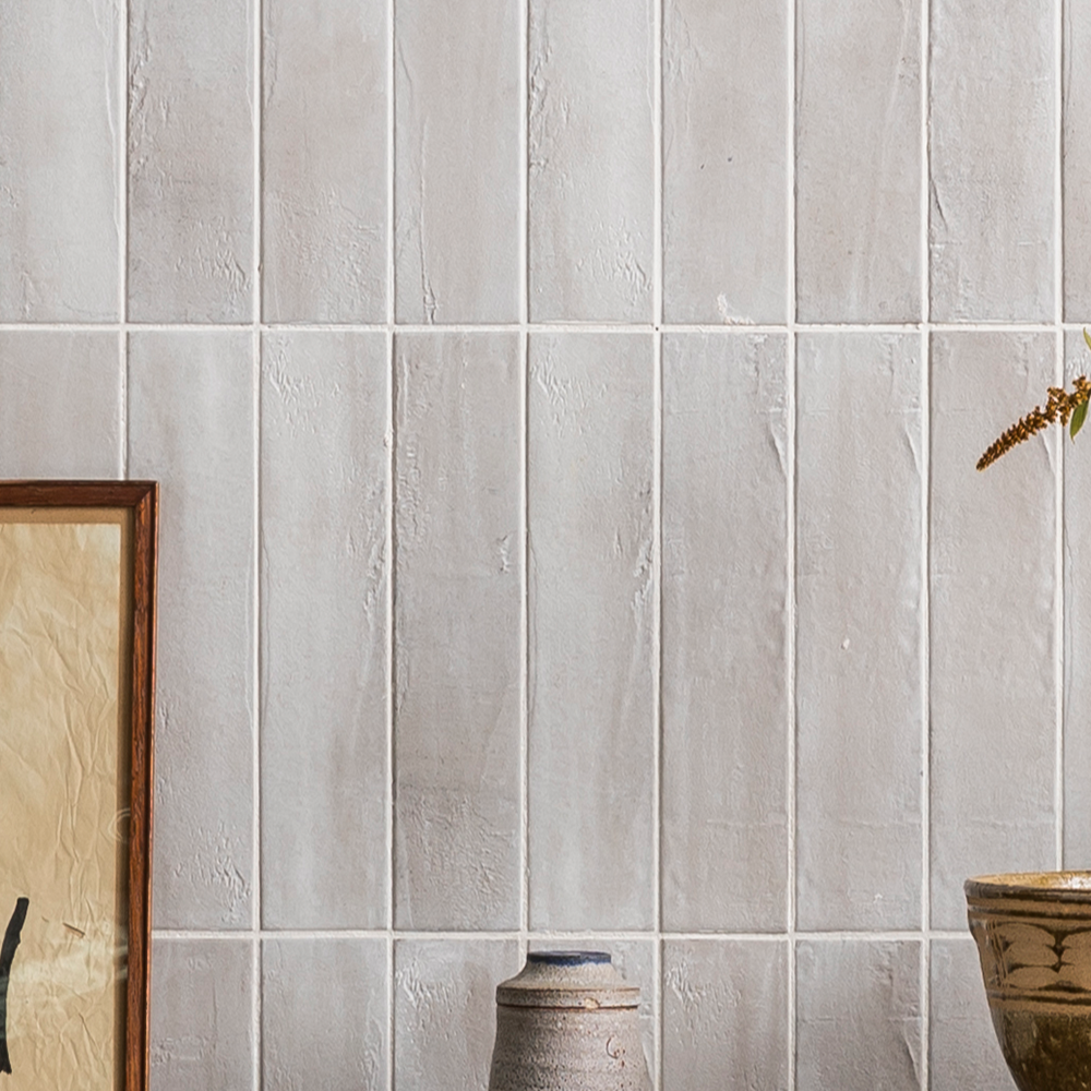 A textured white tiled wall with vertical rectangular tiles. In front, partial views of a framed picture, a ceramic vase, and a potted plant with a dry stem are visible.