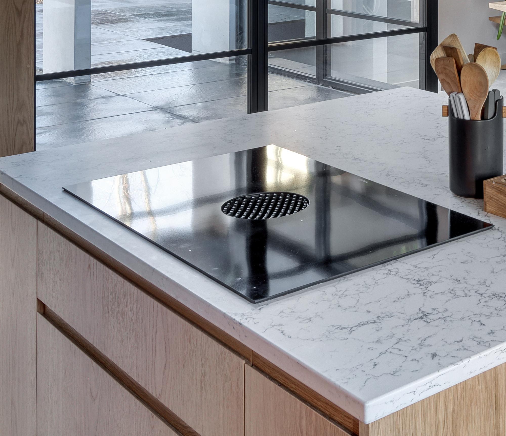A modern kitchen countertop with a sleek black induction cooktop featuring a built-in vent in the center. Wooden cabinets and a container with kitchen utensils are visible beside the cooktop.