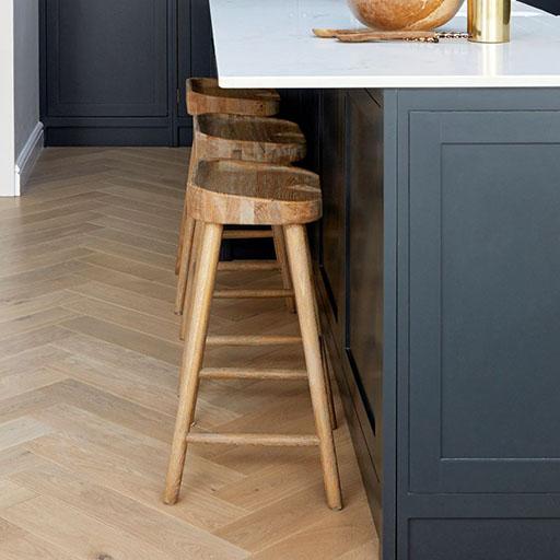 Three wooden bar stools are lined up beside a dark kitchen island with a white countertop, on a light wood herringbone floor. A decorative bowl and gold object sit on the counter.