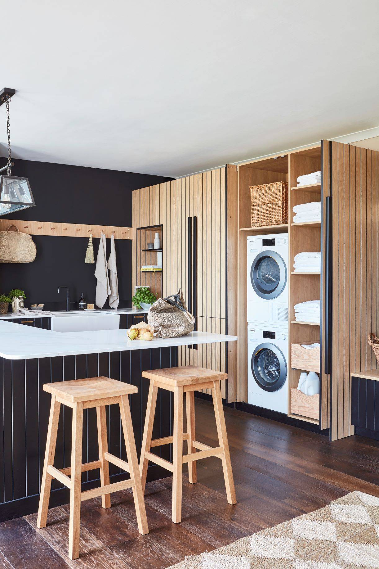 Modern laundry room with wood paneling and dark accents. Features a stacked washer and dryer, open shelves with towels, a basket, pull-out drawers, and a wall-mounted coat rack with an orange apron. Dark wood floors add warmth to the space.