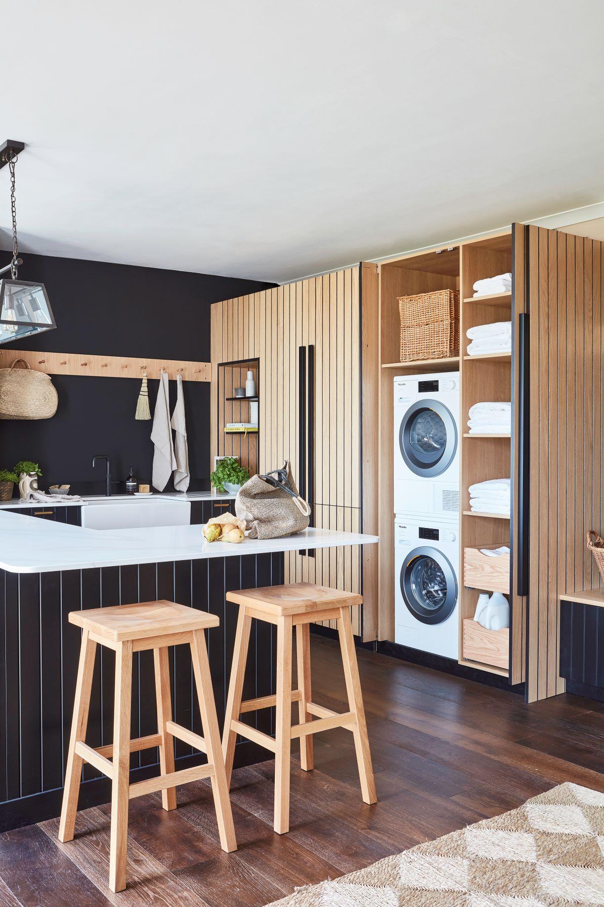 Modern laundry room with wood paneling and dark accents. Features a stacked washer and dryer, open shelves with towels, a basket, pull-out drawers, and a wall-mounted coat rack with an orange apron. Dark wood floors add warmth to the space.