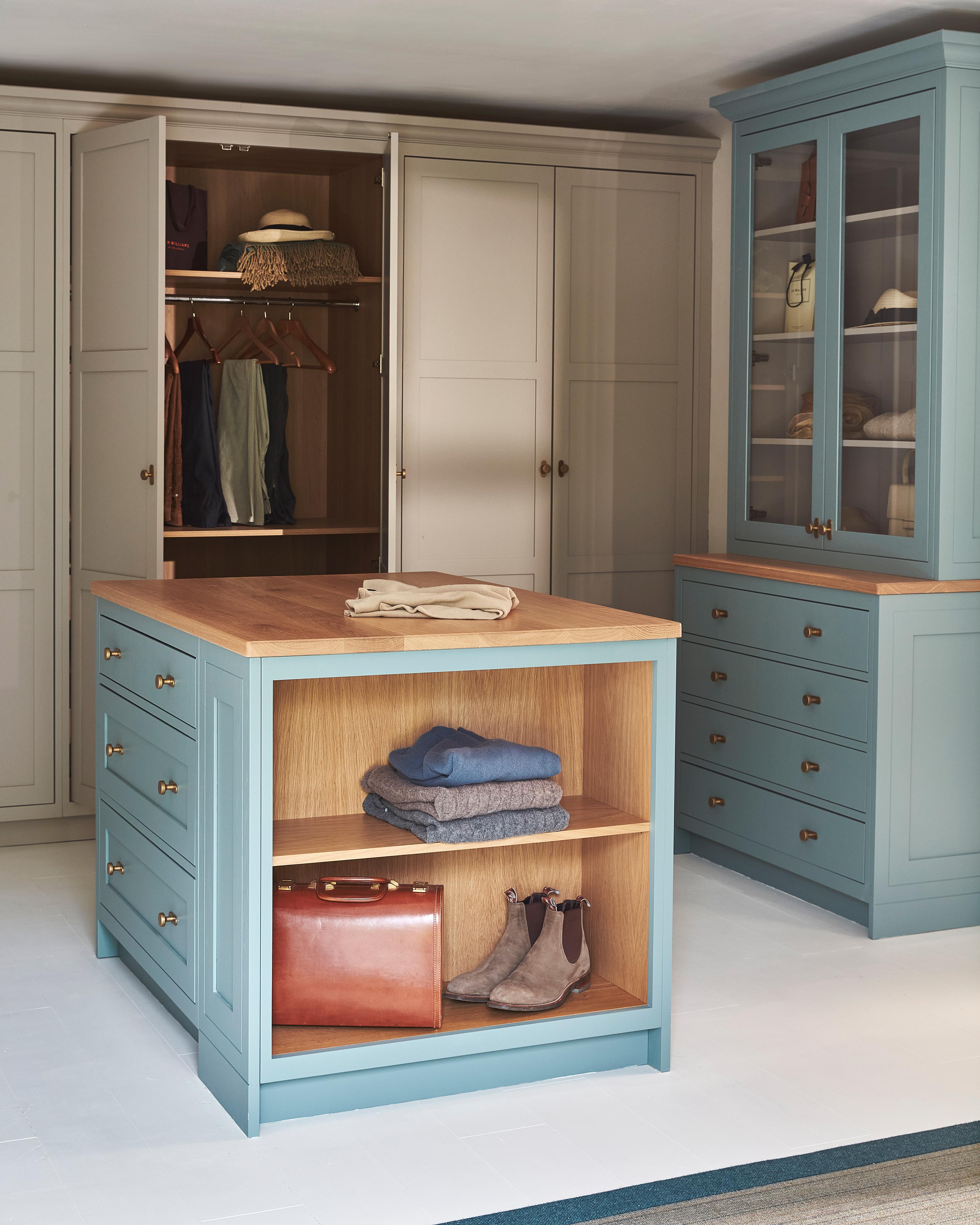 A spacious, organized walk-in closet with light blue cabinets, wood accents, open shelves displaying folded clothes, boots, and a leather bag, and hanging clothes in open wardrobes. A folded sweater rests on the central island.