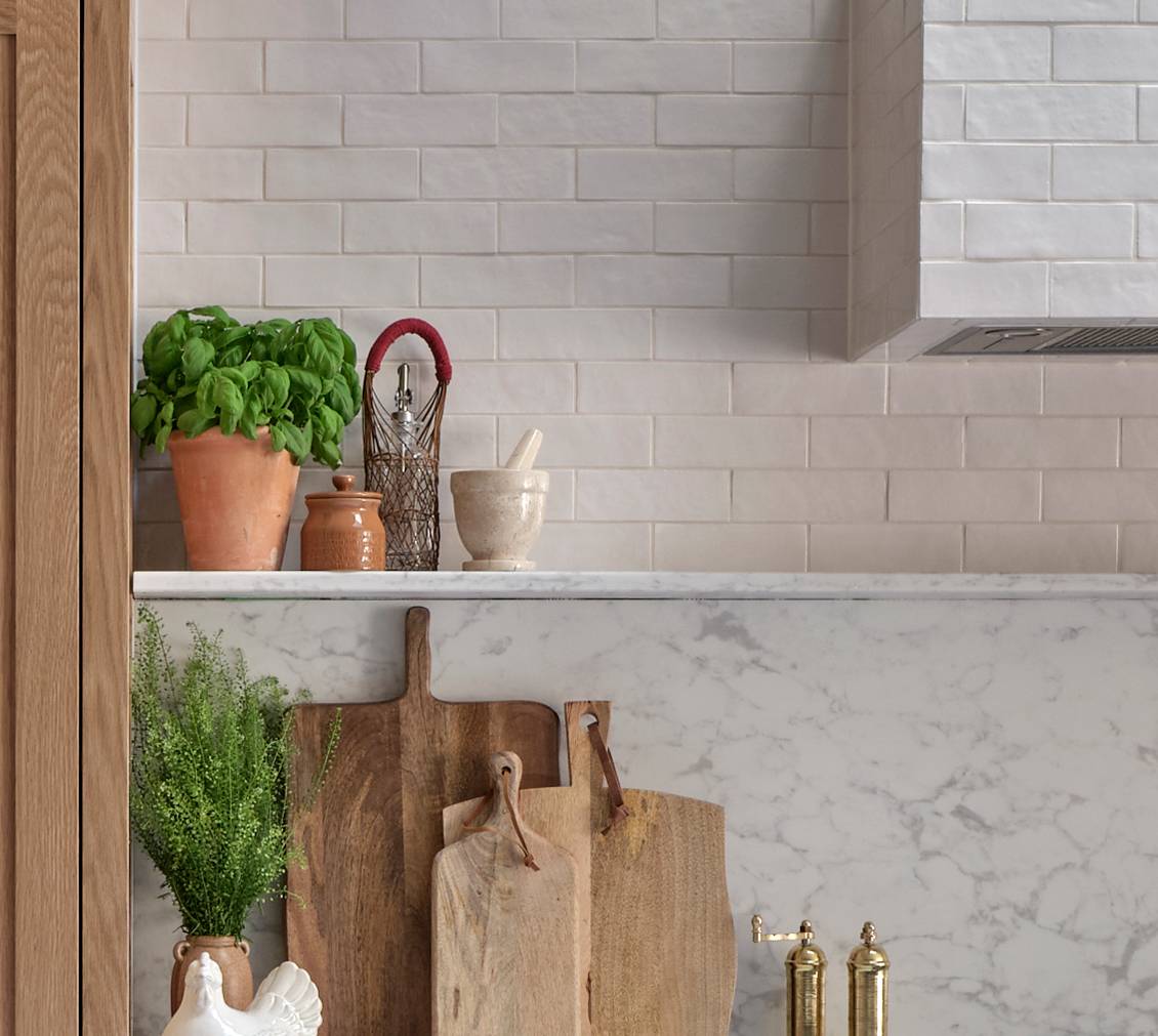 A kitchen counter with a marble backsplash, wooden cutting boards, potted herbs, a ceramic rooster, a mortar and pestle, and kitchen utensils in a jar against a white tile wall.