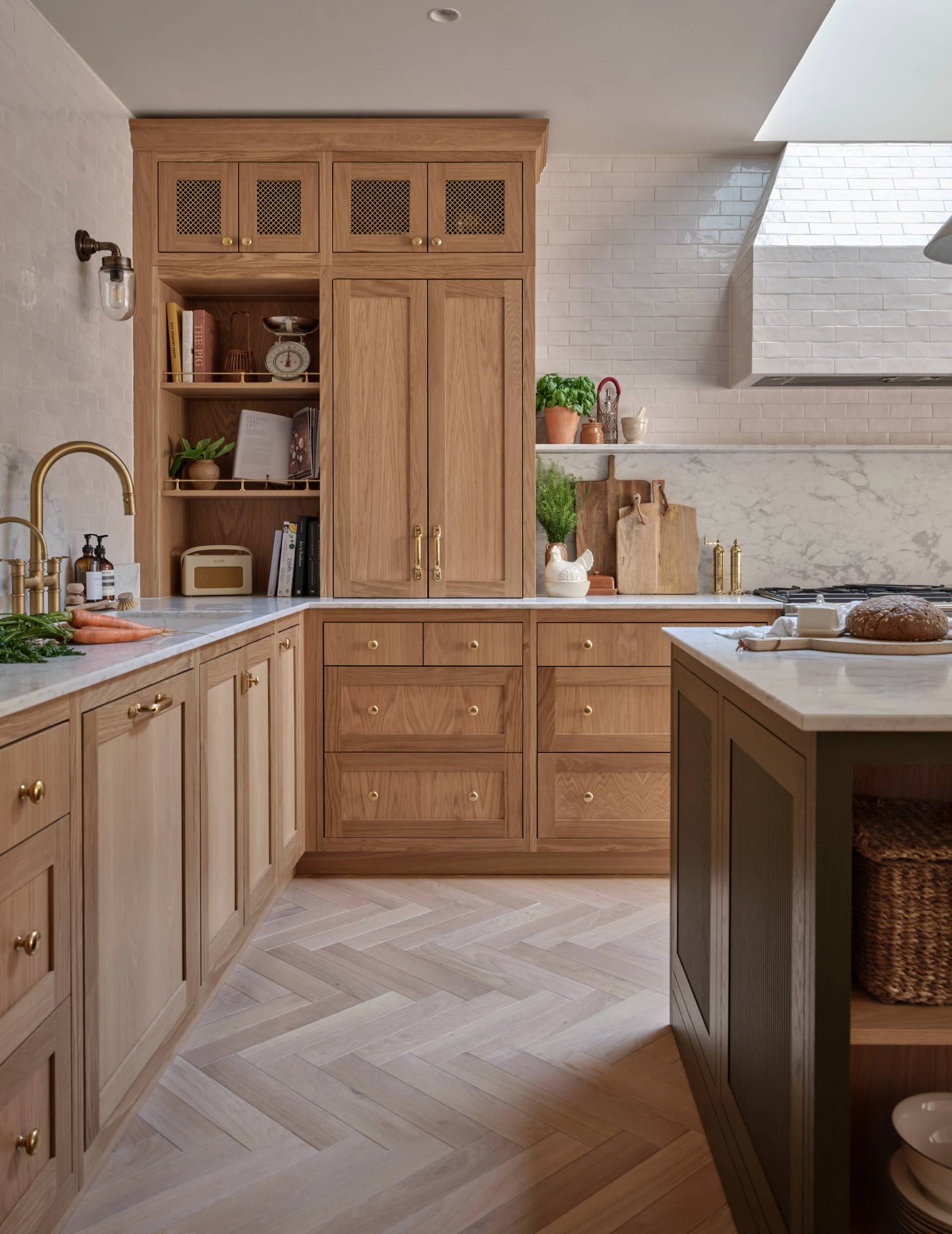 A modern kitchen with light wood cabinets, brass hardware, white marble countertops, herringbone wood floors, open shelves with plants and books, and a skylight providing natural light.