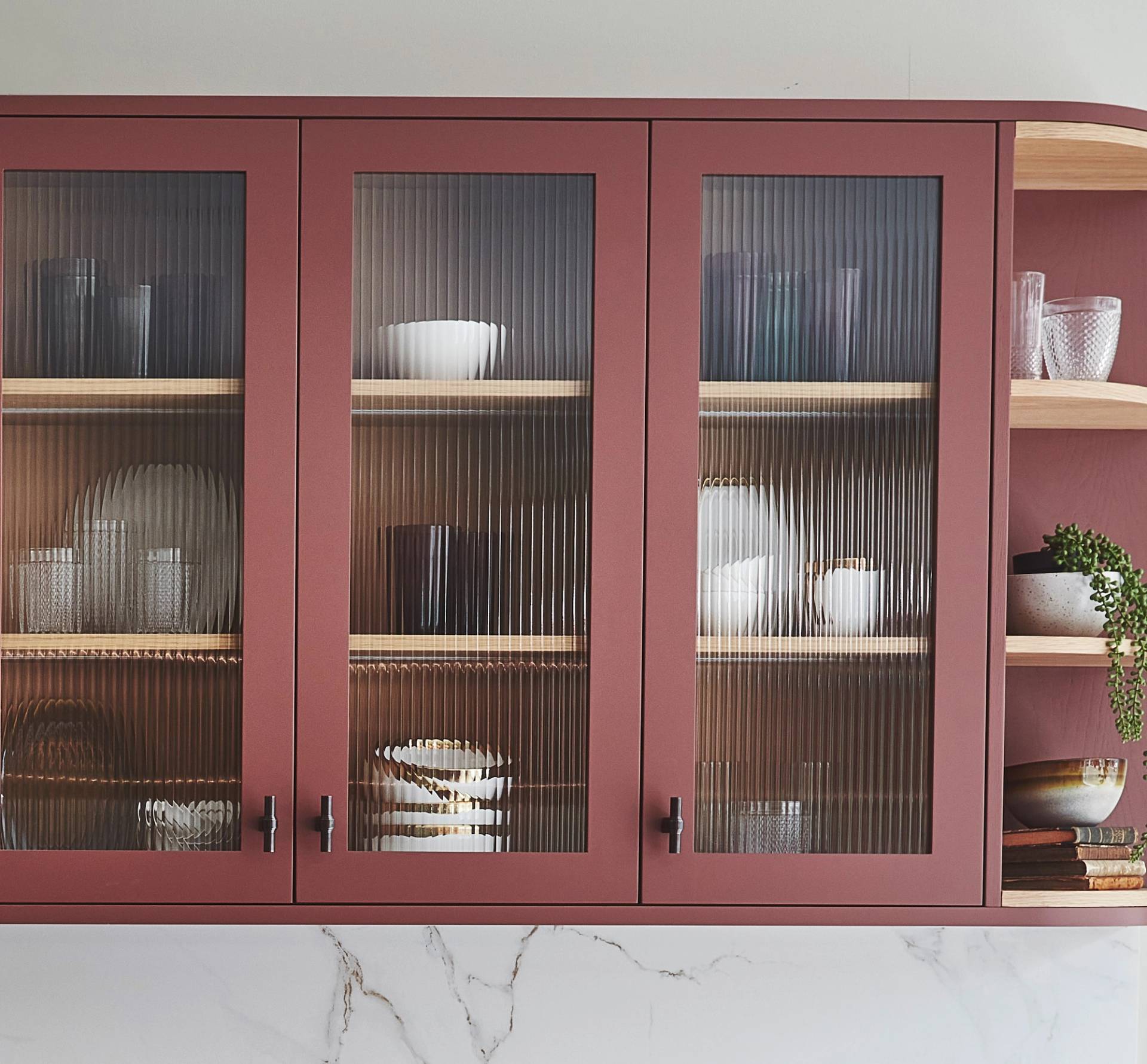A kitchen cabinet with three glass doors, painted in muted red, reveals neatly arranged dishes and glasses. Two open shelves on the side display plants and additional dishware. The cabinet is set against a light wall with a marble countertop beneath.