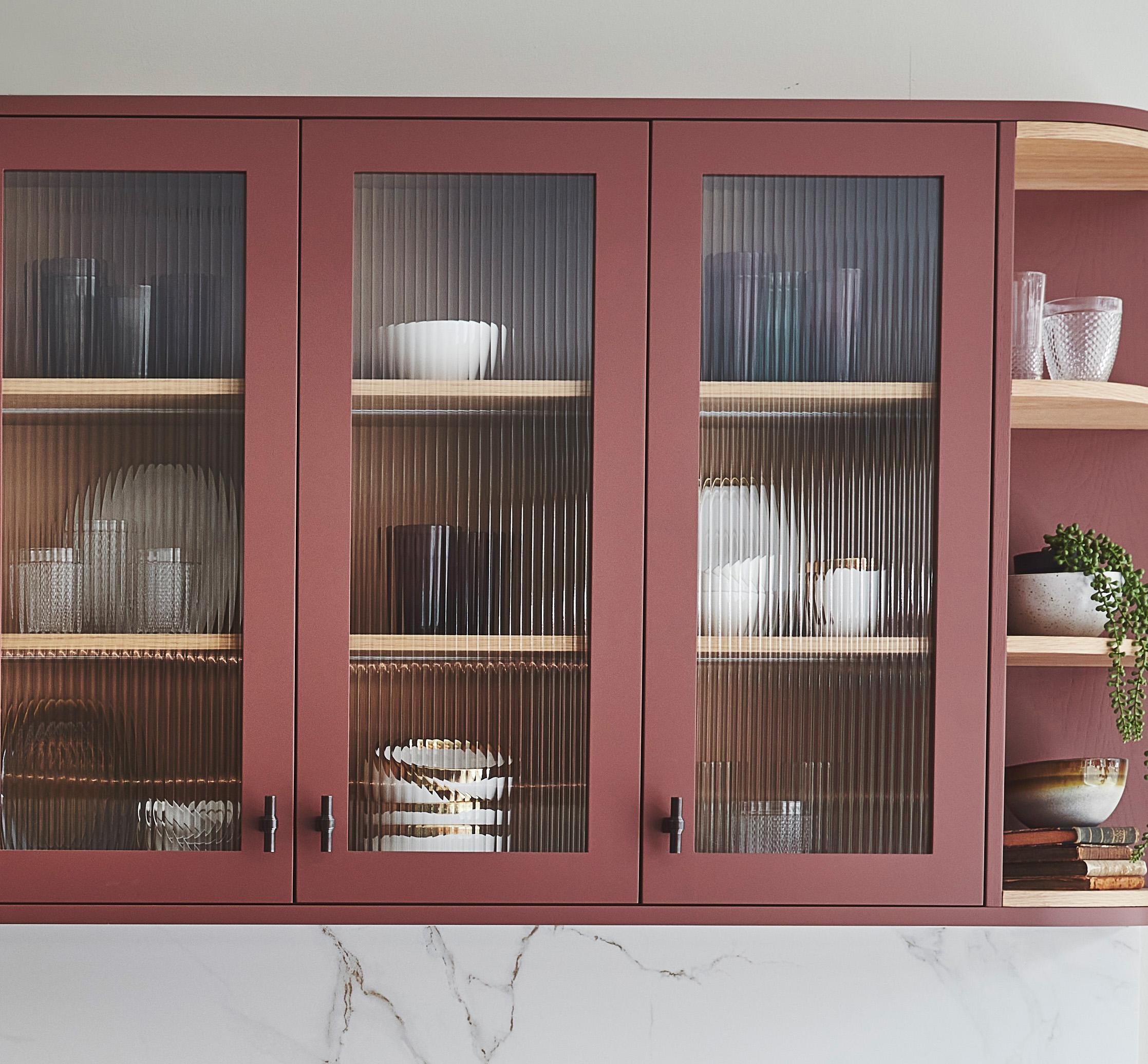 A kitchen cabinet with three glass doors, painted in muted red, reveals neatly arranged dishes and glasses. Two open shelves on the side display plants and additional dishware. The cabinet is set against a light wall with a marble countertop beneath.