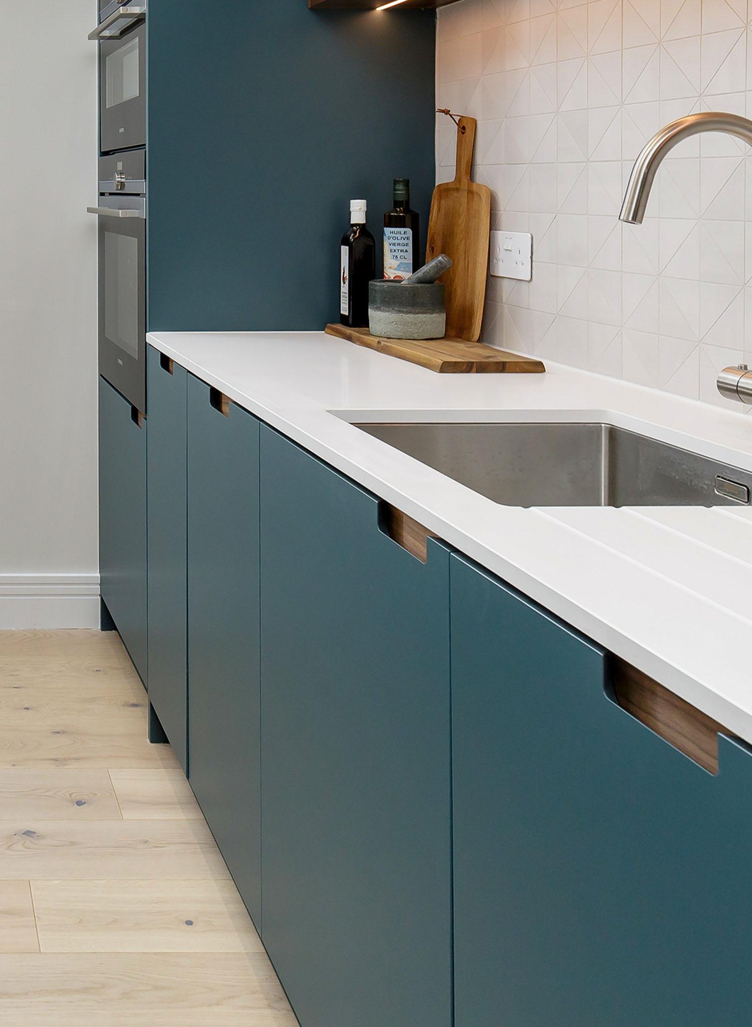 Modern kitchen with teal cabinets, white countertop, built-in oven, stainless steel sink, and geometric tile backsplash. Wooden utensils, bottles, and a cutting board are arranged near the wall.