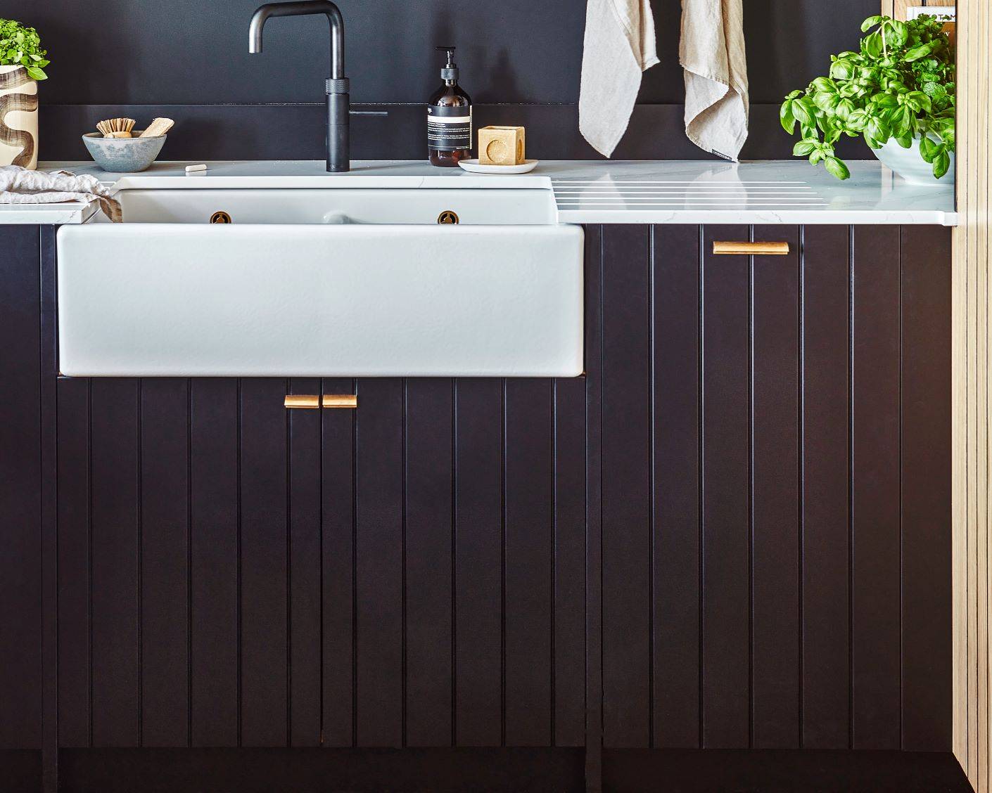 A modern kitchen features a white farmhouse sink with a black faucet, dark vertical panel cabinets, gold handles, a striped countertop, a potted basil plant, towels, and kitchen essentials neatly arranged.