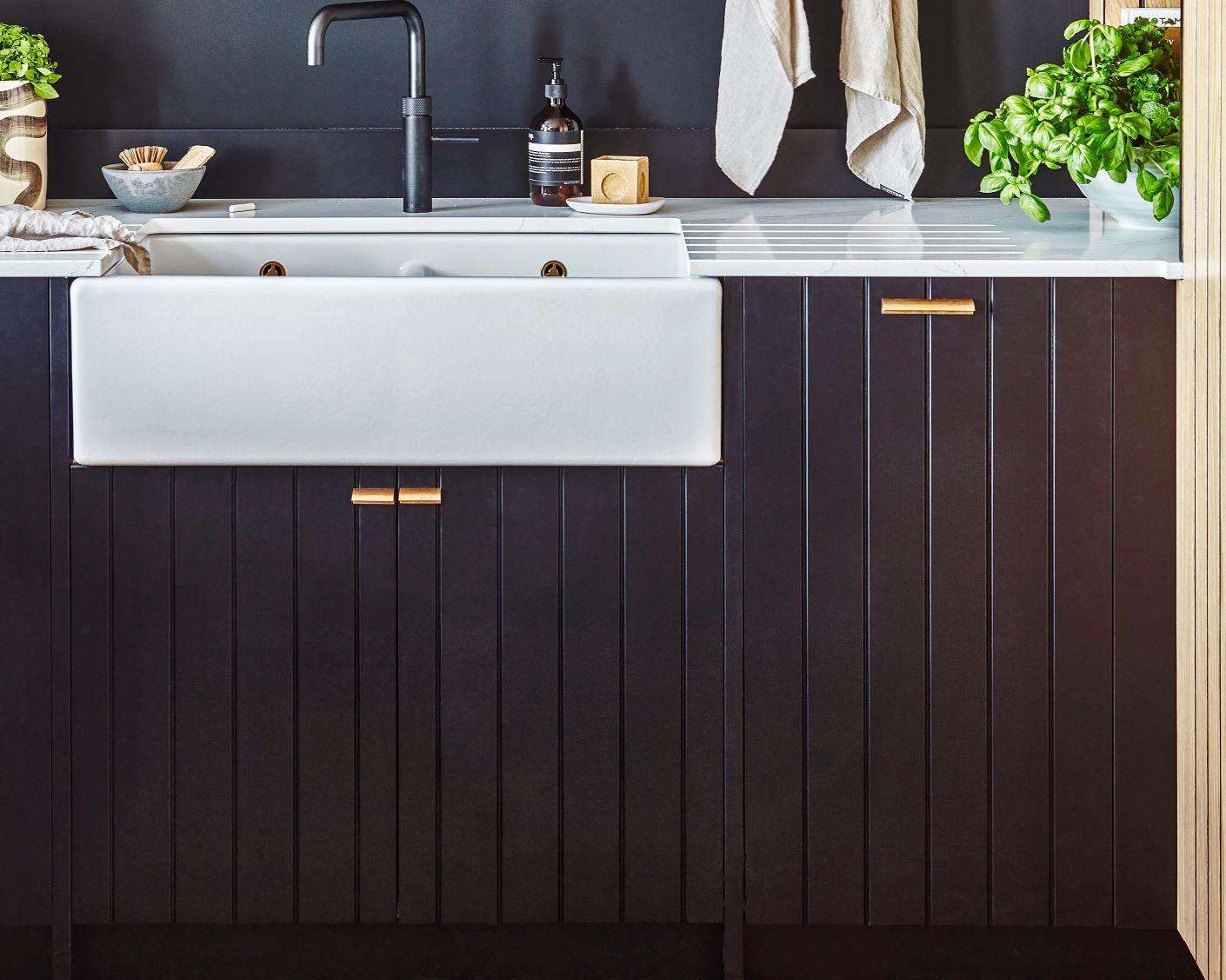 A modern kitchen features a white farmhouse sink with a black faucet, dark vertical panel cabinets, gold handles, a striped countertop, a potted basil plant, towels, and kitchen essentials neatly arranged.