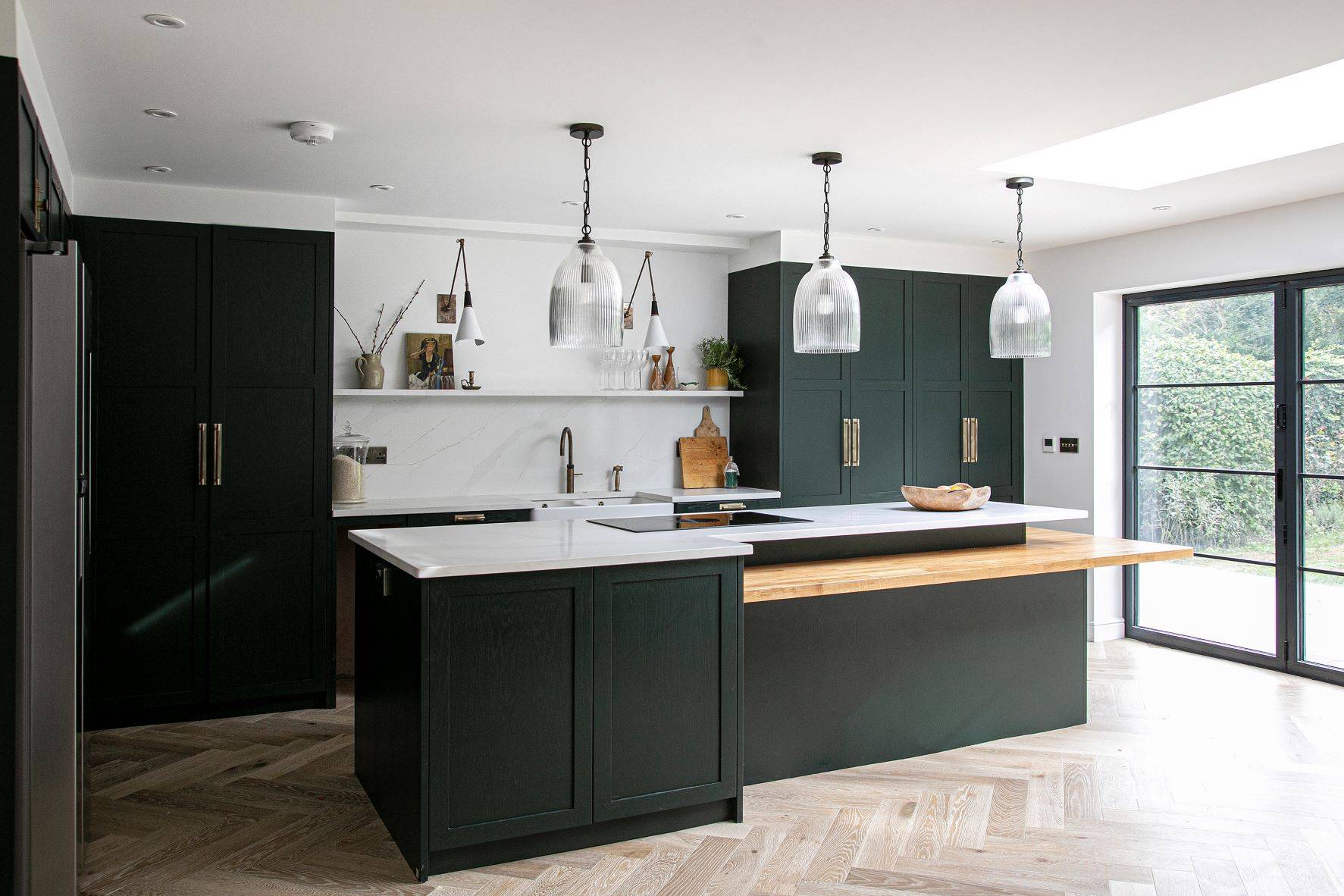 A modern kitchen with dark green cabinets and white countertops. It features a central island with wooden accents, three pendant lights, open shelves with decor, and large windows providing natural light. The floor is a light herringbone pattern.