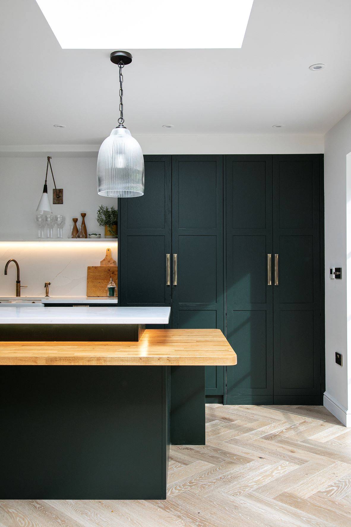 A modern kitchen with dark green cabinets, brass handles, and light wood countertops. The island features a built-in wooden table extension. A large, textured glass pendant light hangs from the ceiling. The floor is light wood, and there's soft under-cabinet lighting.