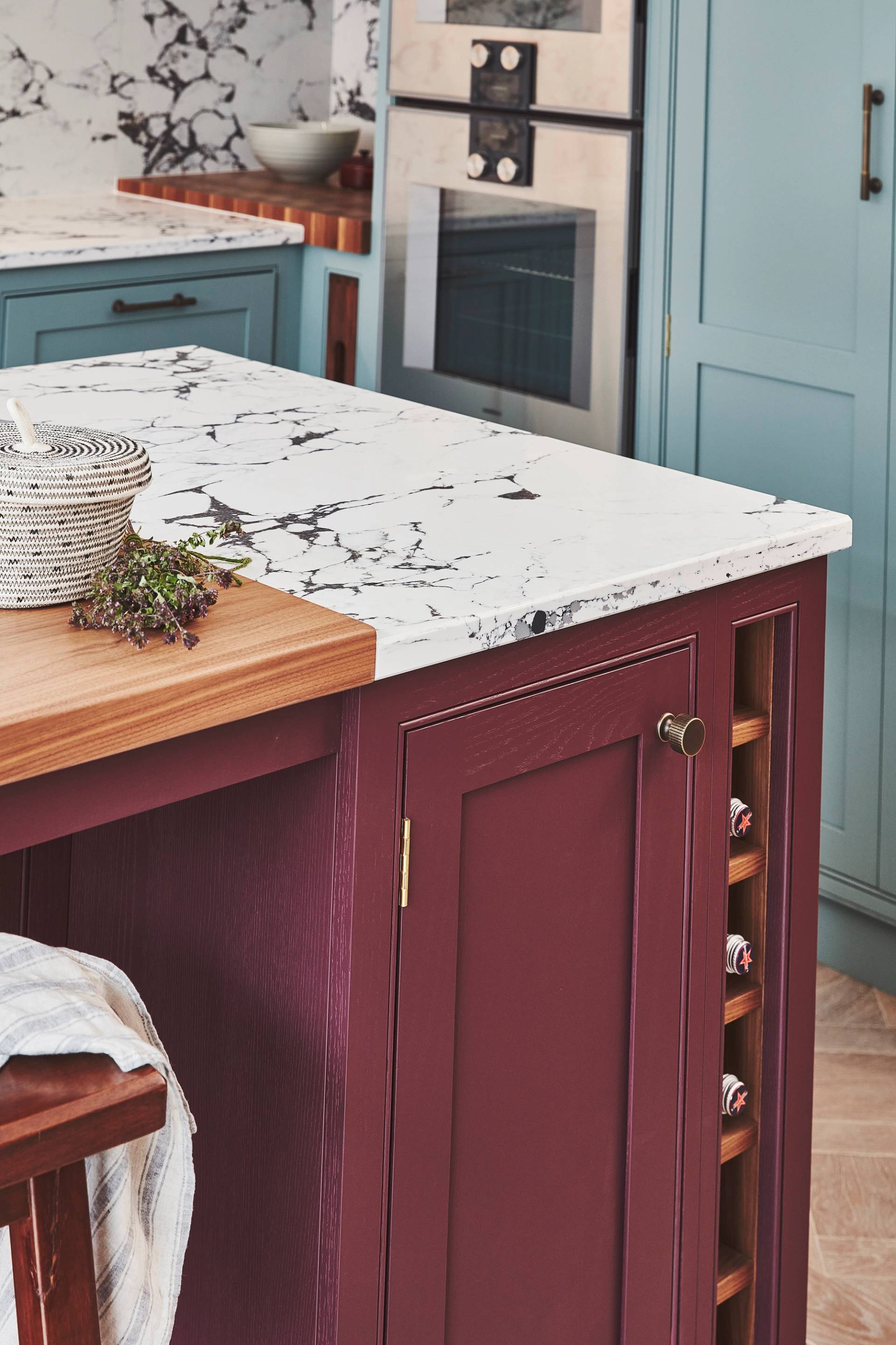 A modern kitchen with a marble countertop, deep burgundy cabinetry, and a built-in wine rack. Blue cabinets and dual ovens are in the background. A wooden cutting board and a basket with herbs are on the counter.
