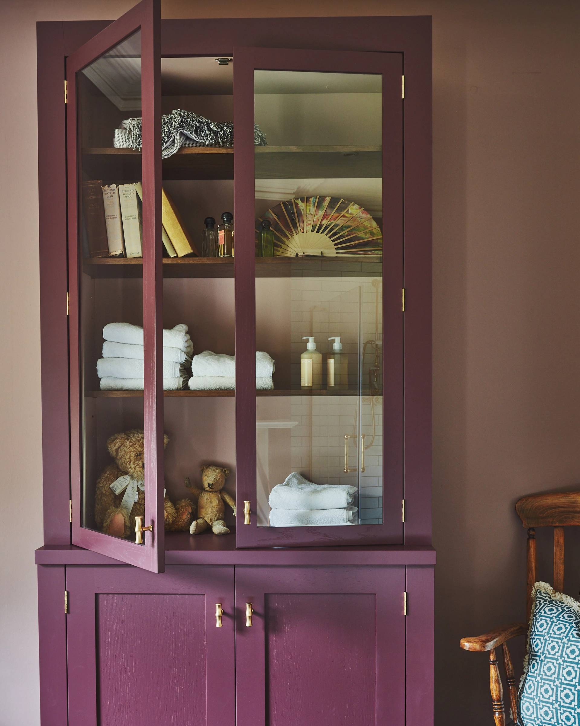 A maroon cabinet with glass doors showcases folded towels, books, two teddy bears, bottles, and decorative items on shelves. A wooden chair with a patterned cushion is partially visible next to it. The setting has a cozy, vintage feel.