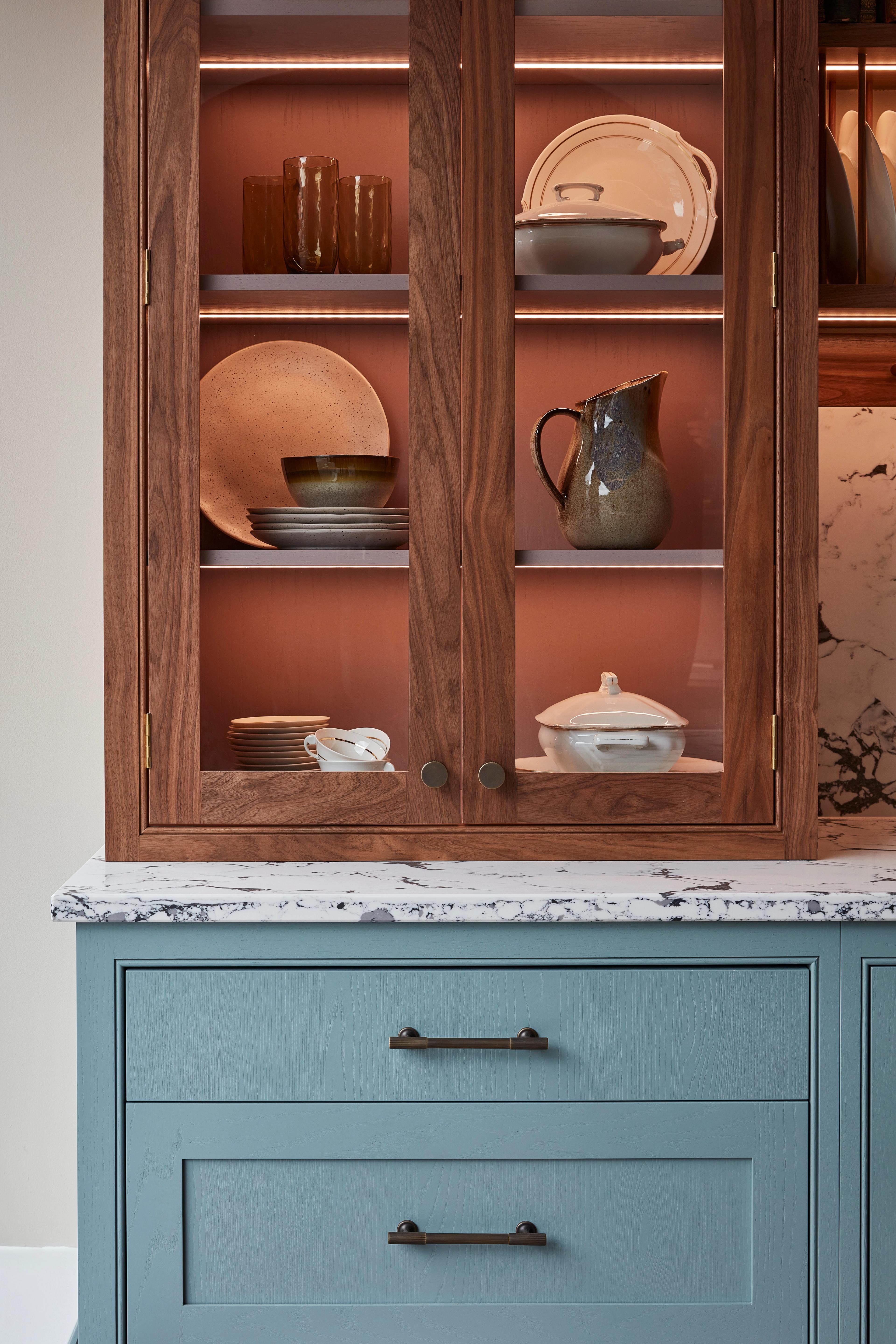A wooden cabinet with glass doors displaying ceramic dishes, bowls, and a jug. Below is a light blue drawer set with a marbled countertop. Warm lighting highlights the textures and tones of the ceramics and wood