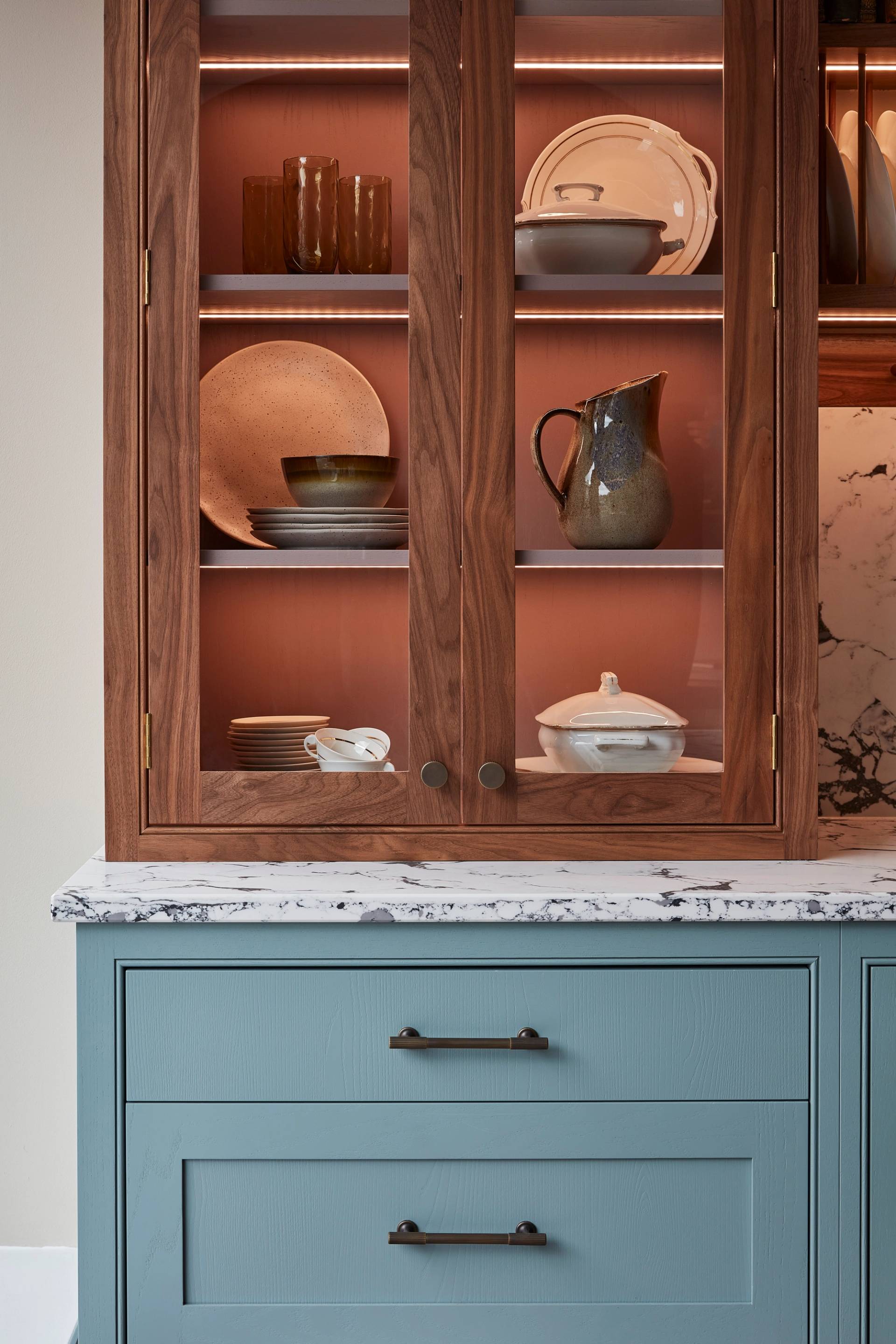 A wooden cabinet with glass doors displaying ceramic dishes, bowls, and a jug. Below is a light blue drawer set with a marbled countertop. Warm lighting highlights the textures and tones of the ceramics and wood