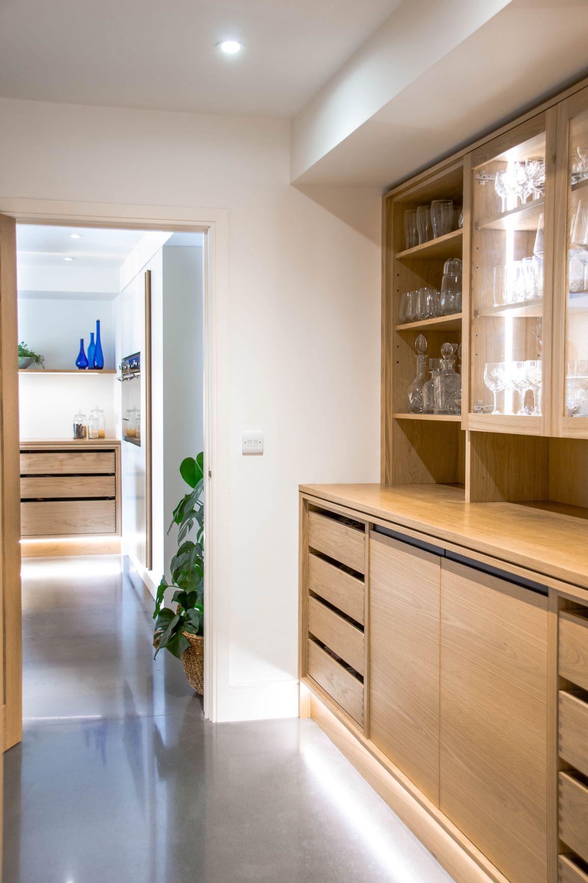 Wooden kitchen cabinets with glass doors display various glassware. Open shelves hold bottles and cups. The countertop is clear, and lower drawers provide additional storage space. The overall look is clean and organized.