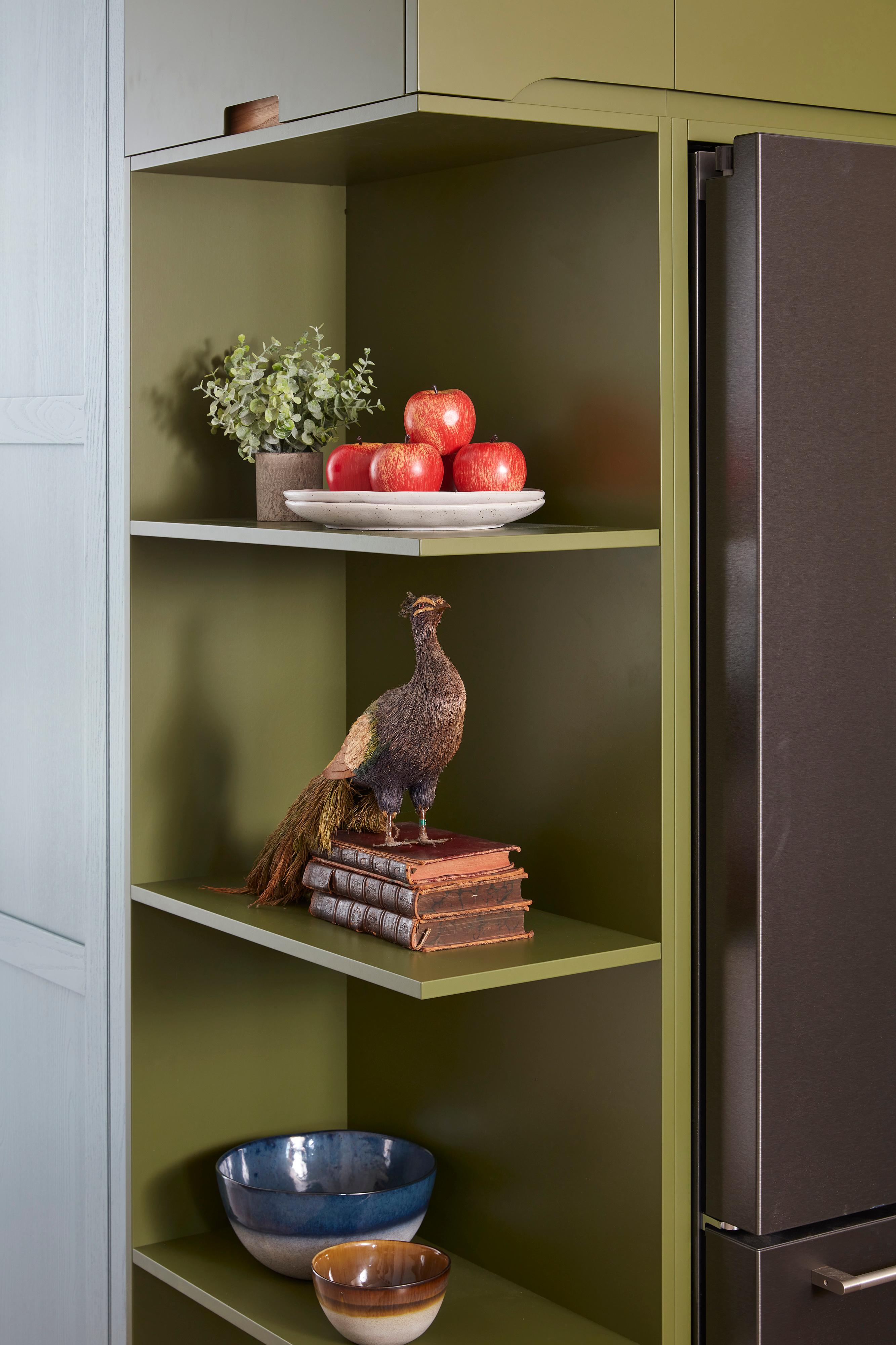 A kitchen shelf with green walls holds a decorative bird on top of a stack of books, a plate of red apples with greenery, and two bowls. A refrigerator is partially visible on the right.