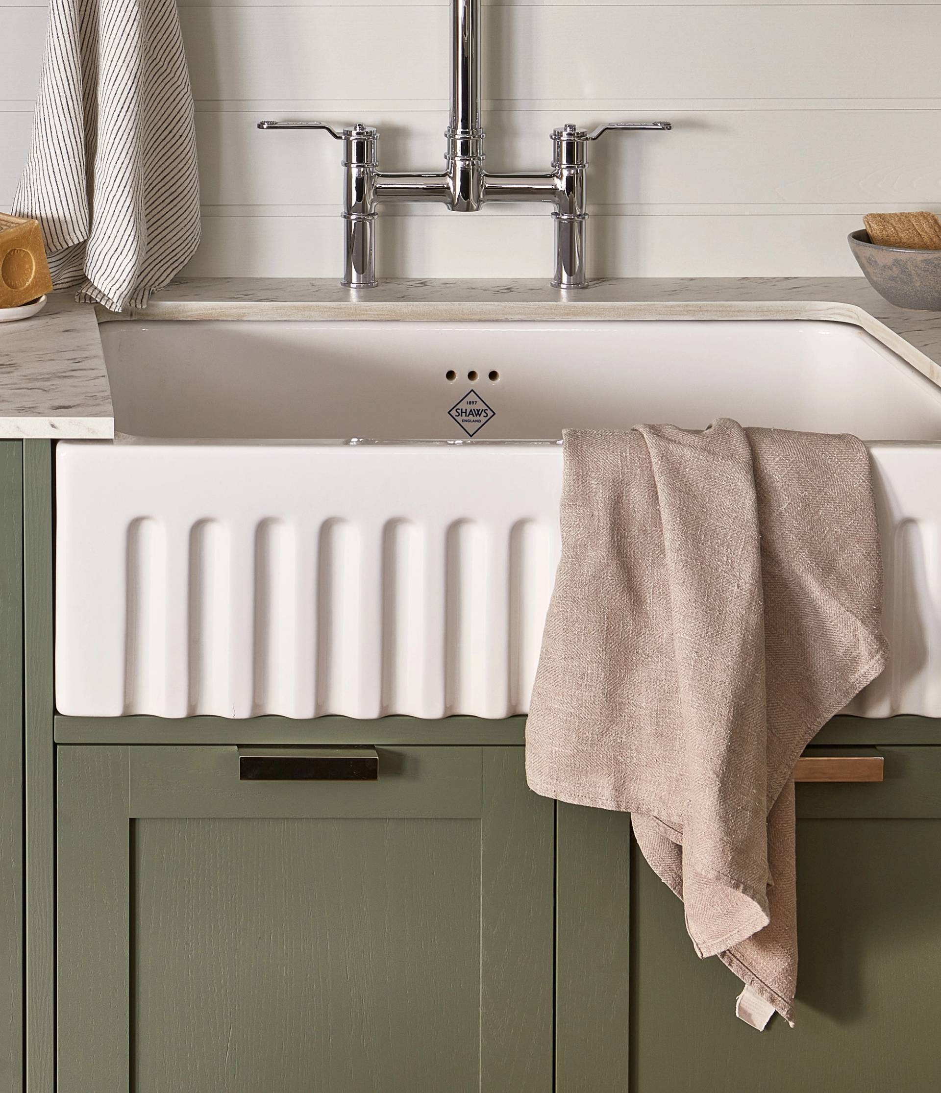 A kitchen sink with a chrome faucet is set in a white countertop. A beige towel hangs over the front of the white, ridged basin. The cabinet below is olive green with a marble-like accent to the left. A striped cloth hangs nearby.