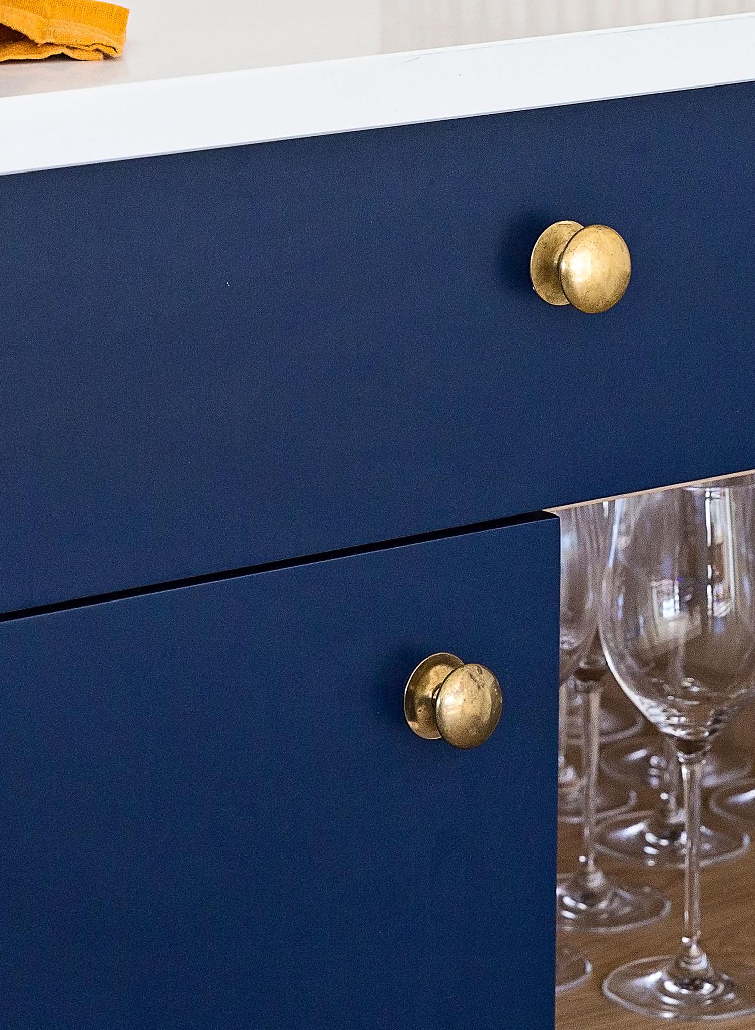 A close-up of blue kitchen cabinets with round brass knobs, a white countertop above, and several wine glasses stored inside the cabinet below.