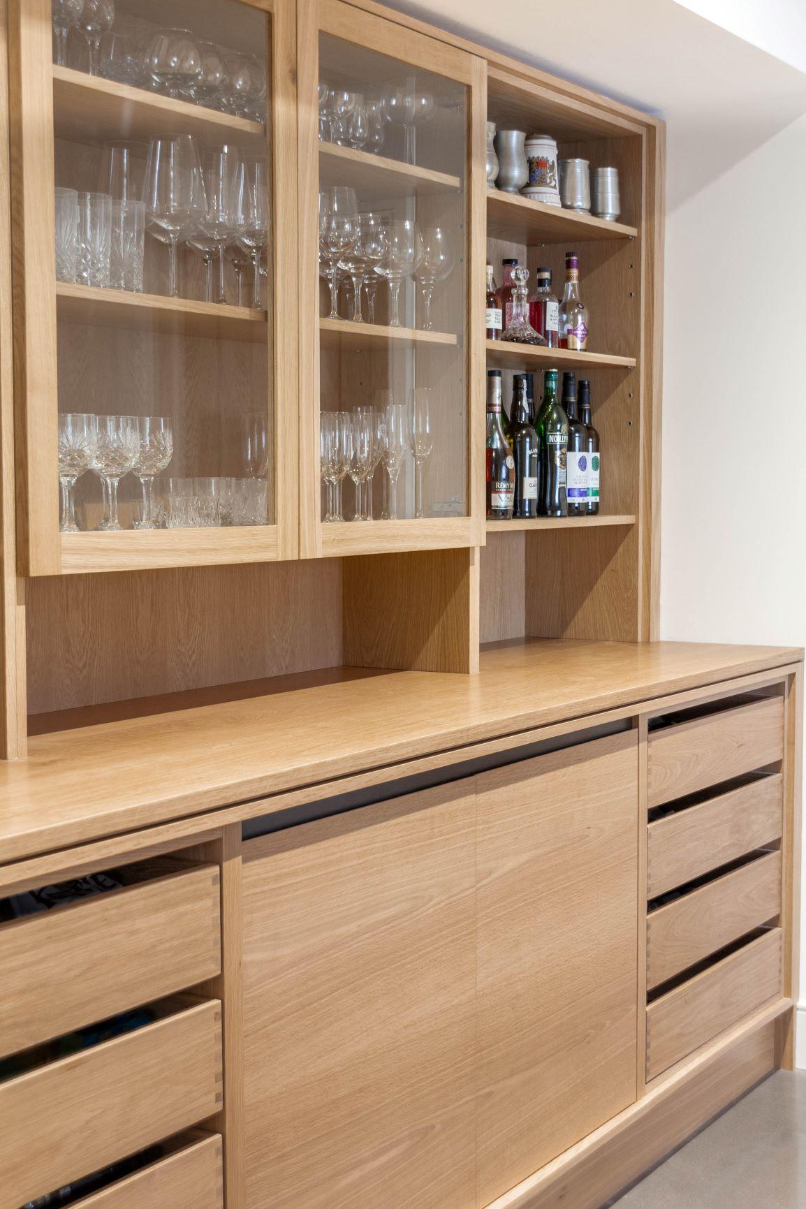 Wooden kitchen cabinets with glass doors display various glassware. Open shelves hold bottles and cups. The countertop is clear, and lower drawers provide additional storage space. The overall look is clean and organized.