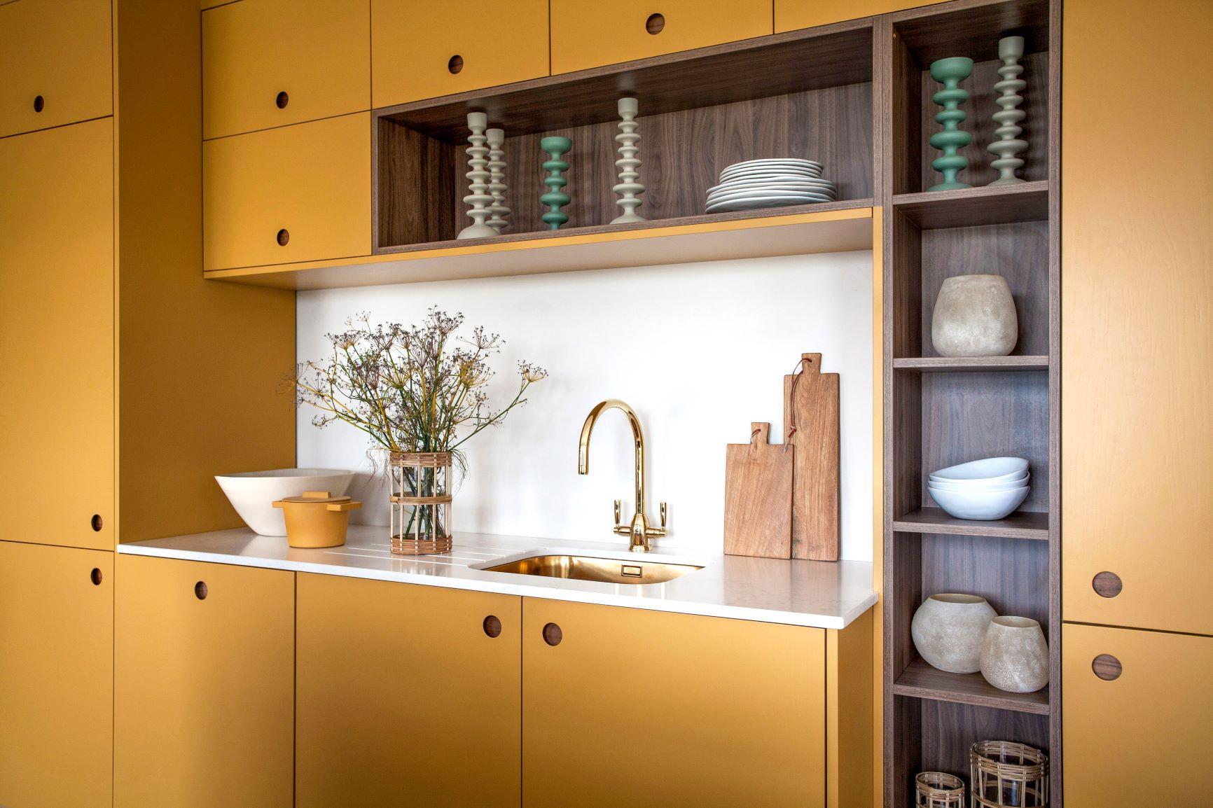 A modern kitchen with mustard yellow cabinets, a white countertop, and a gold faucet. Shelves display decorative vases, candlesticks, and stacked plates. A vase with flowers and wooden cutting boards add accents.