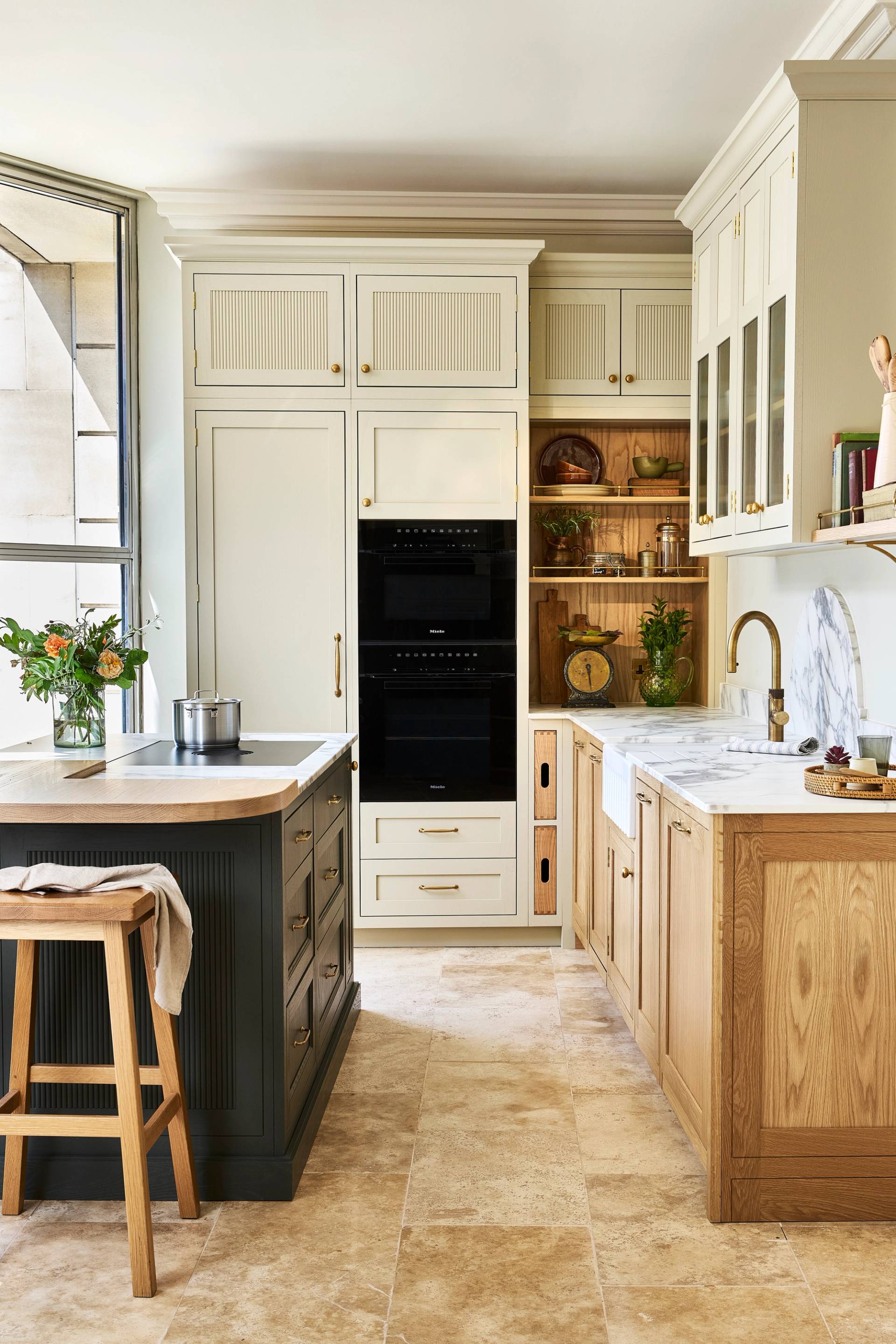 Bright, modern kitchen with pale blue cabinets, a farmhouse sink, white walls, open shelves holding dishes and plants, a window with a sheer white blind, and a dark herringbone-patterned floor.