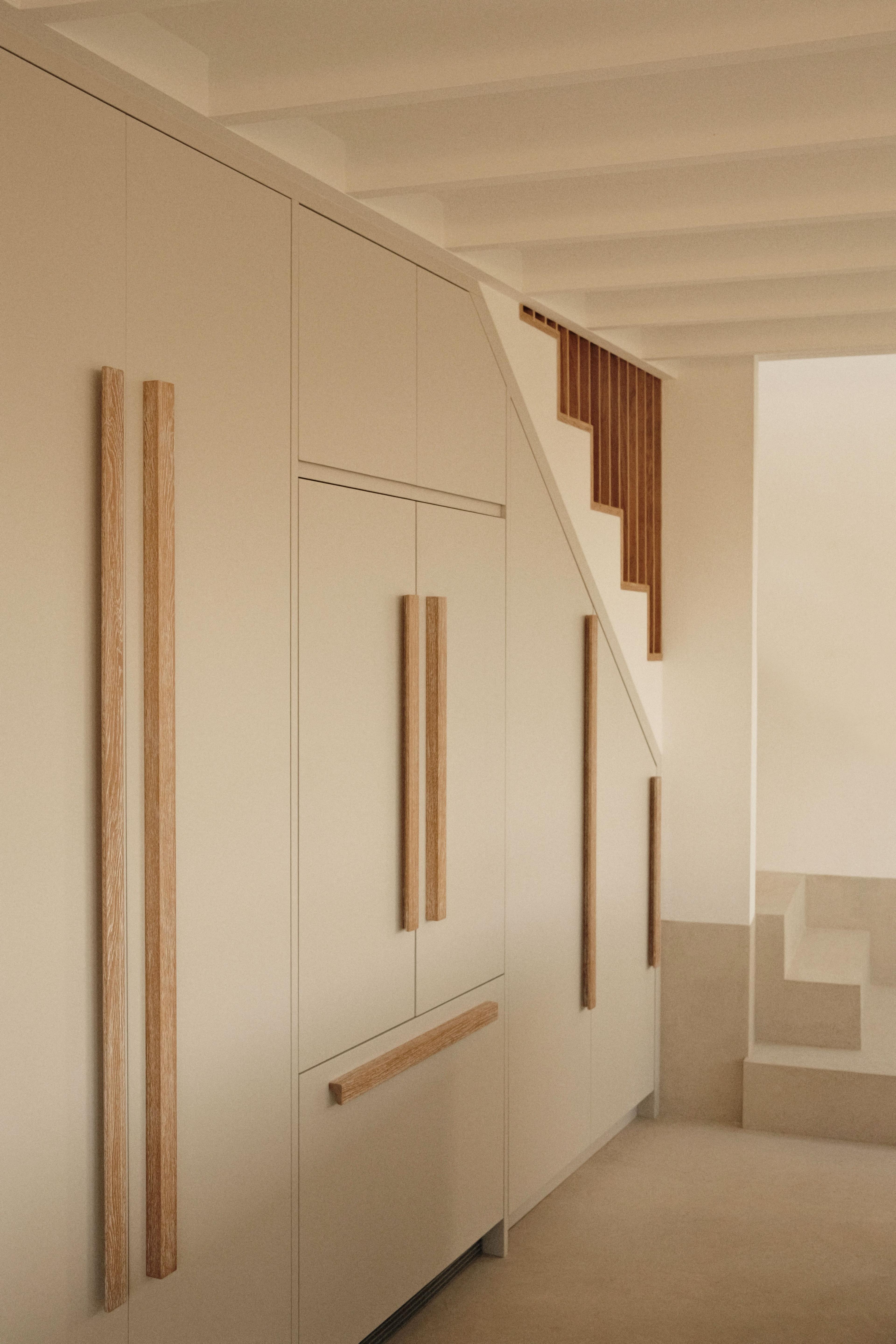Minimalist interior with beige built-in cabinets featuring long, vertical wooden handles, located under a staircase with wooden railings and light-colored steps. The space has a clean, modern design.