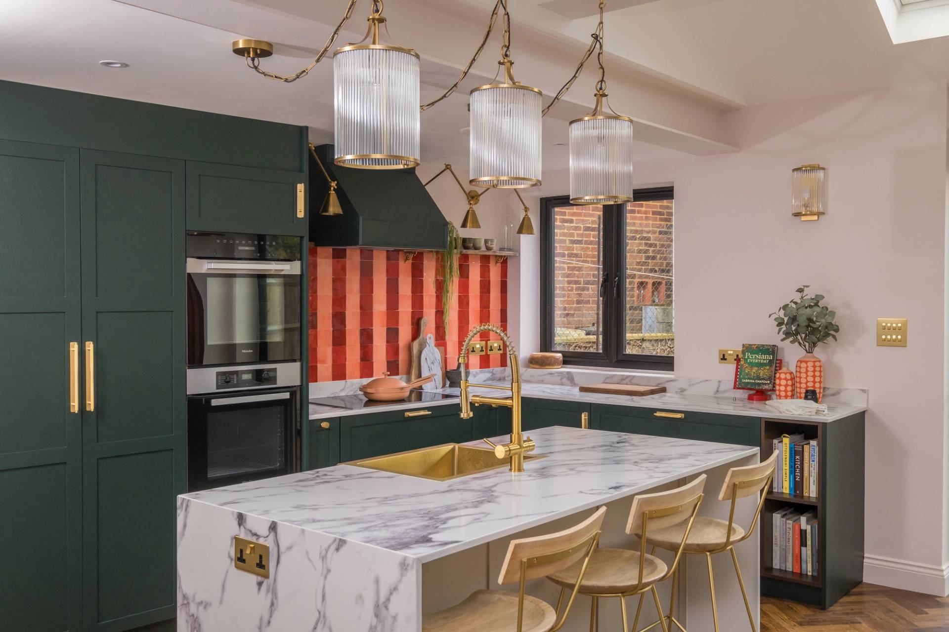 A modern kitchen featuring a marble island with a gold faucet and three beige stools. The kitchen has dark green cabinets, a red checkered backsplash, and stainless steel appliances. Three ribbed pendant lights hang above the island.