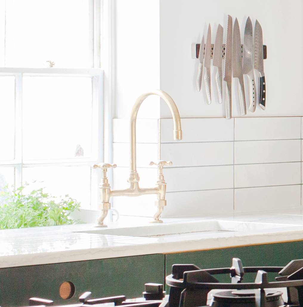 A bright kitchen with a brass faucet, a white countertop, a gas stove in the foreground, and a magnetic knife rack holding several knives on a white-tiled wall. Sunlight streams through a window with a green plant nearby.