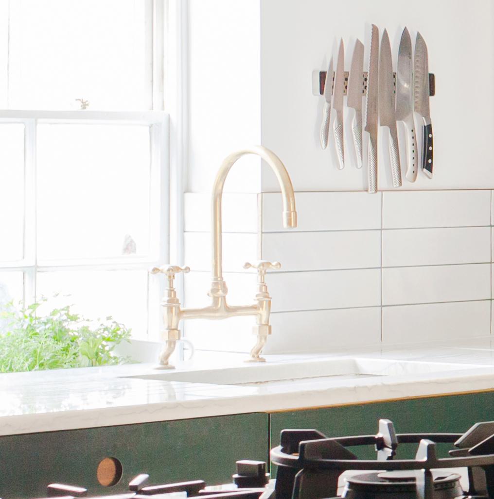 A bright kitchen with a brass faucet, a white countertop, a gas stove in the foreground, and a magnetic knife rack holding several knives on a white-tiled wall. Sunlight streams through a window with a green plant nearby.