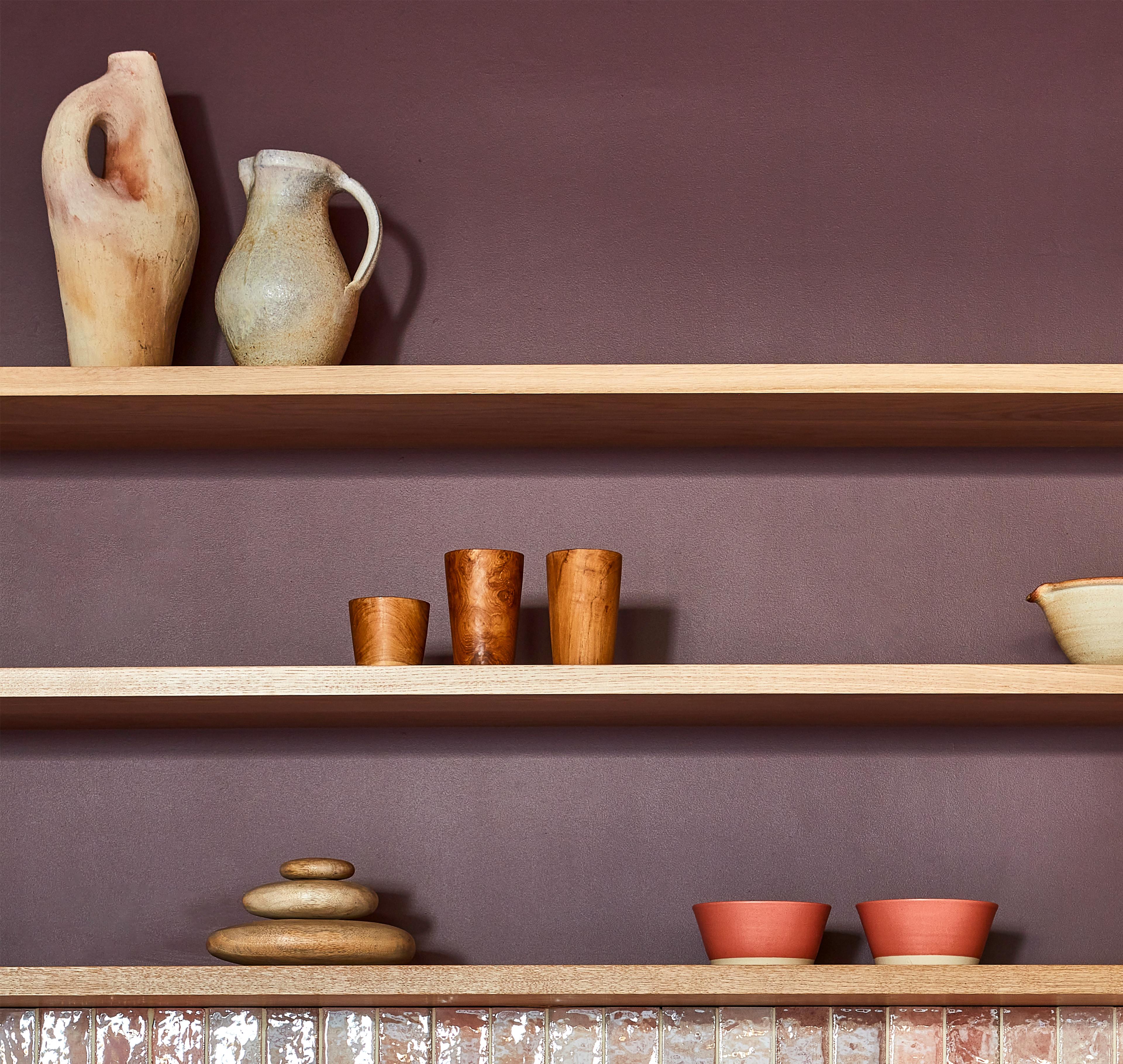 Wooden shelves on a maroon wall display various ceramic items: a tall abstract vase, a pitcher, two wooden cups, a stack of smooth stones, and two small bowls. A row of translucent tile pieces lines the bottom shelf.