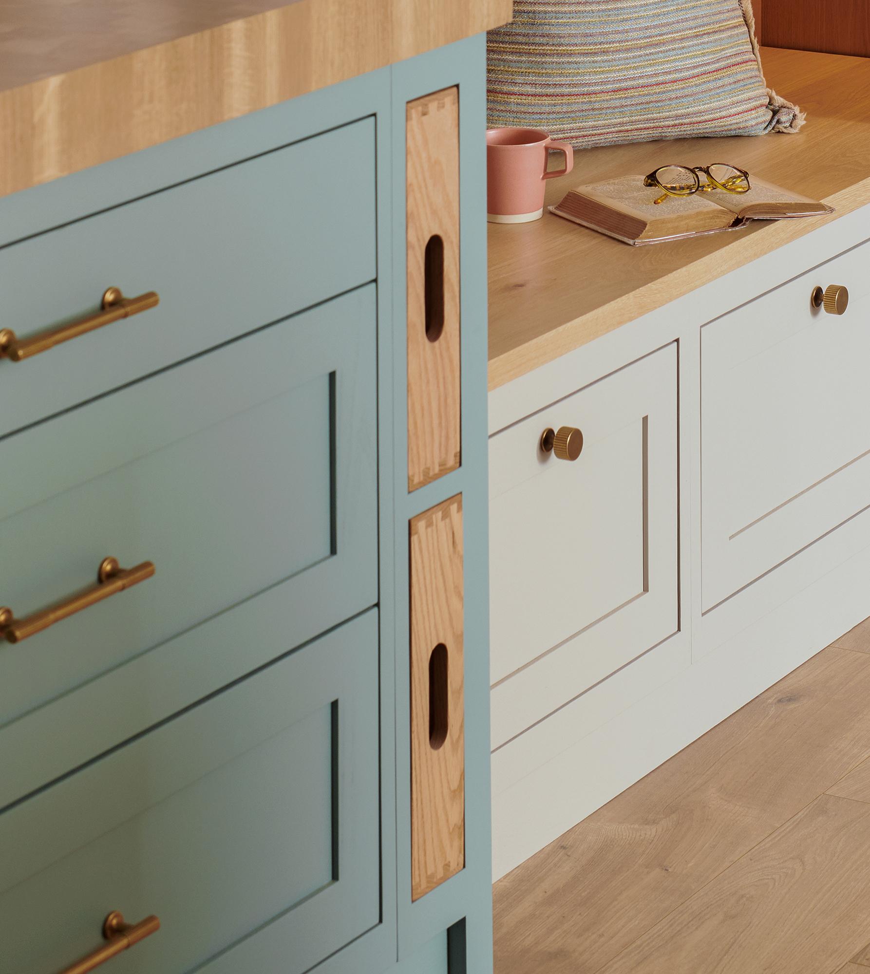 A cozy corner with a light blue drawer unit, light wood countertop, and white cabinets. A colorful cushion, book, glasses, and pink mug sit on the adjacent bench. Warm wooden floor completes the inviting space.