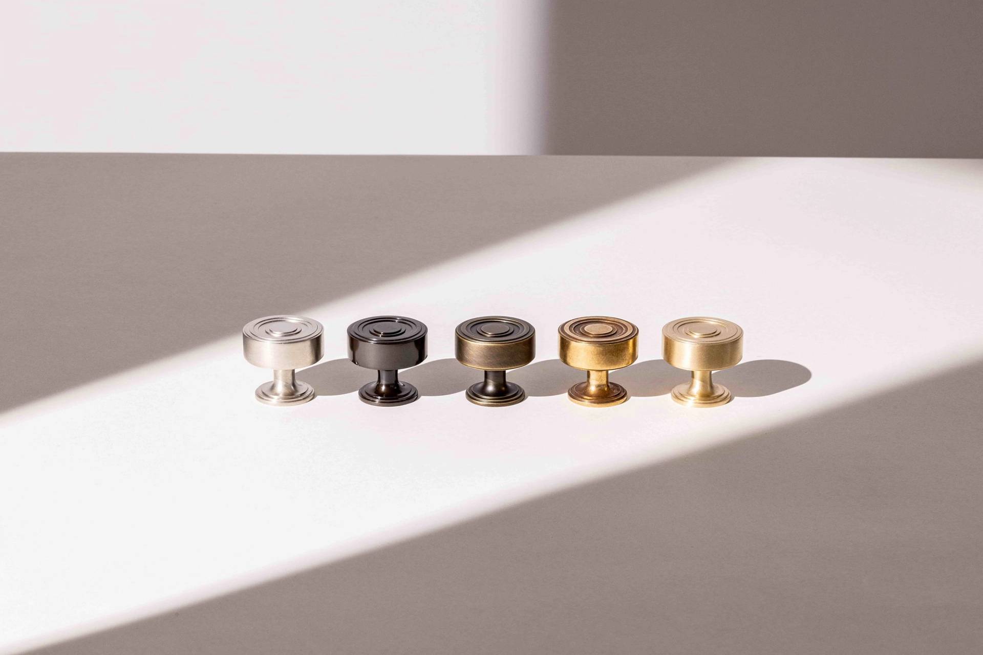 Five metal knobs are aligned on a flat, well-lit surface. The knobs are arranged in color order from left to right: silver, silver-black, black, bronze, and gold. A diagonal shadow cuts across the surface, partially shading the knobs.