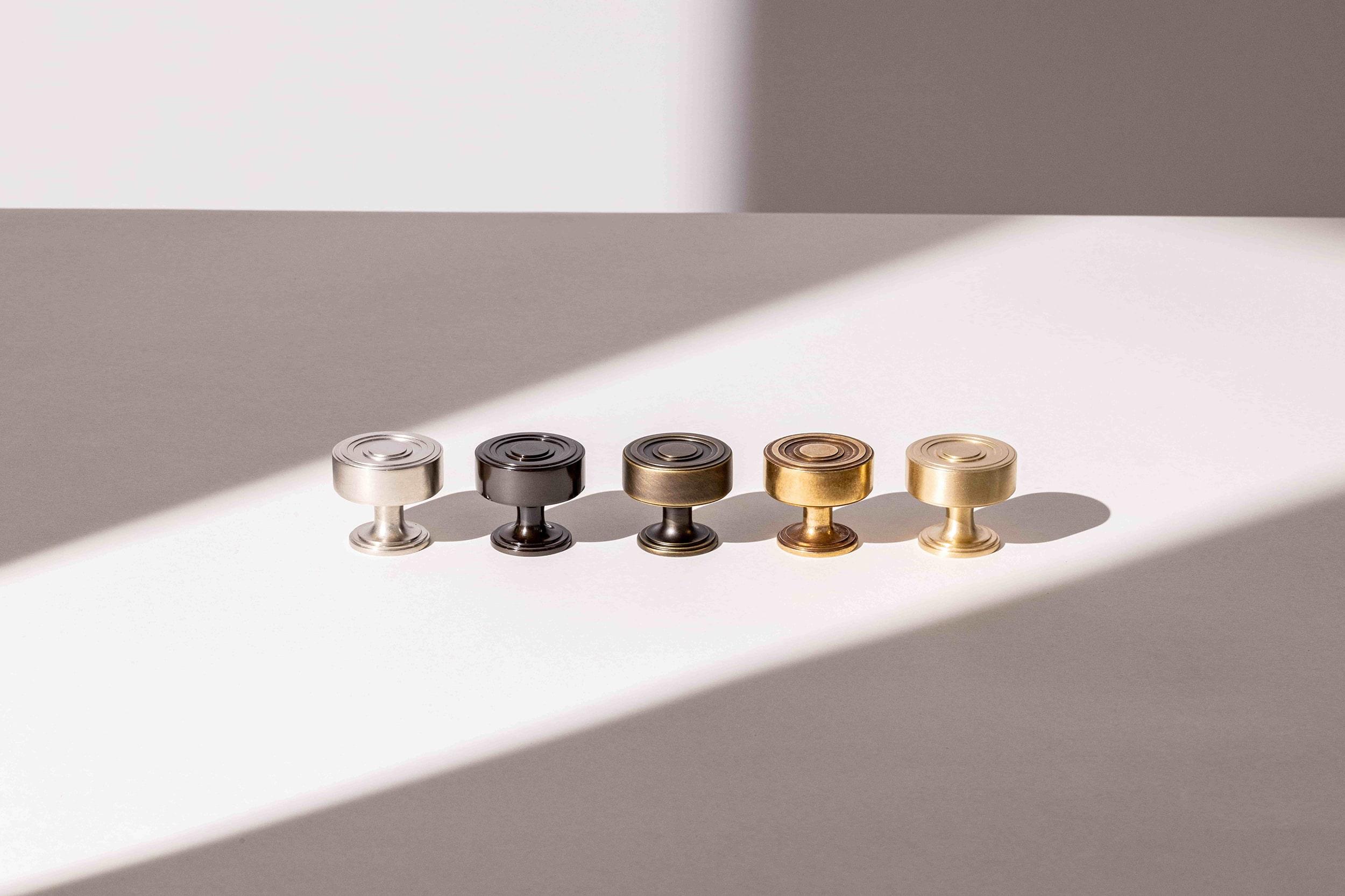 Five metal knobs are aligned on a flat, well-lit surface. The knobs are arranged in color order from left to right: silver, silver-black, black, bronze, and gold. A diagonal shadow cuts across the surface, partially shading the knobs.