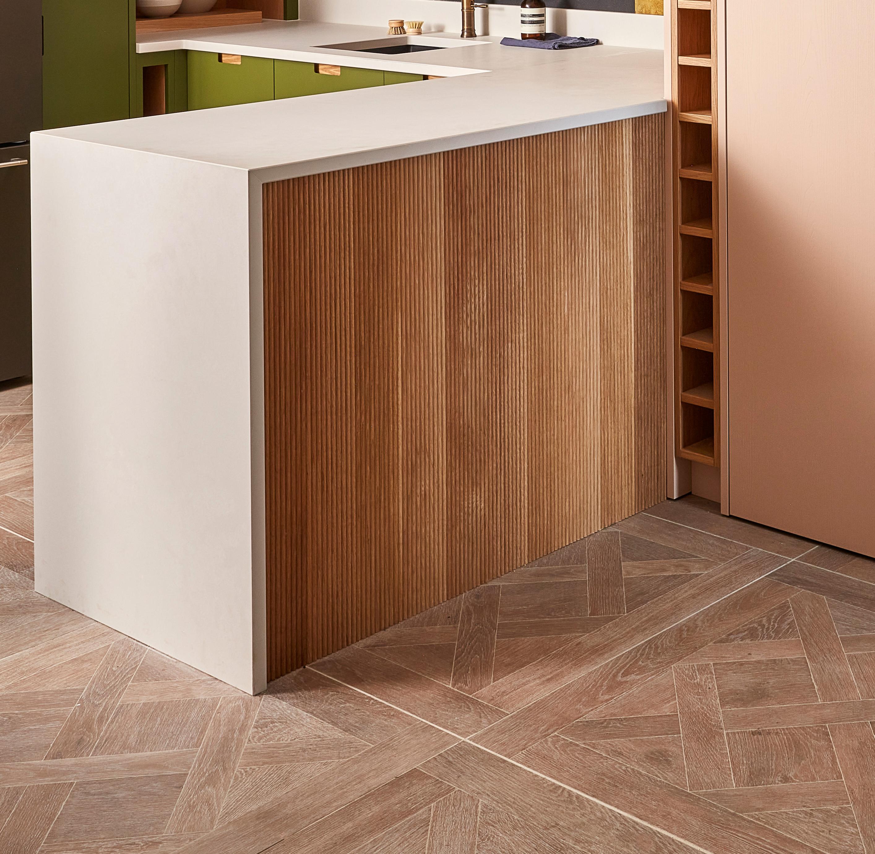 A modern kitchen corner featuring a white countertop, vertical wood paneling on the breakfast bar, geometric wood-patterned floor tiles, green cabinets, and a built-in shelving unit on the right.
