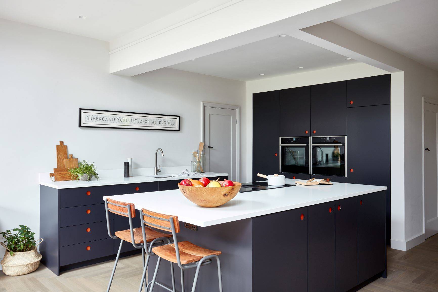 Modern kitchen with dark cabinets and white countertops. A wood bowl with fruit sits on the island, flanked by two wooden chairs. Wall art reads 