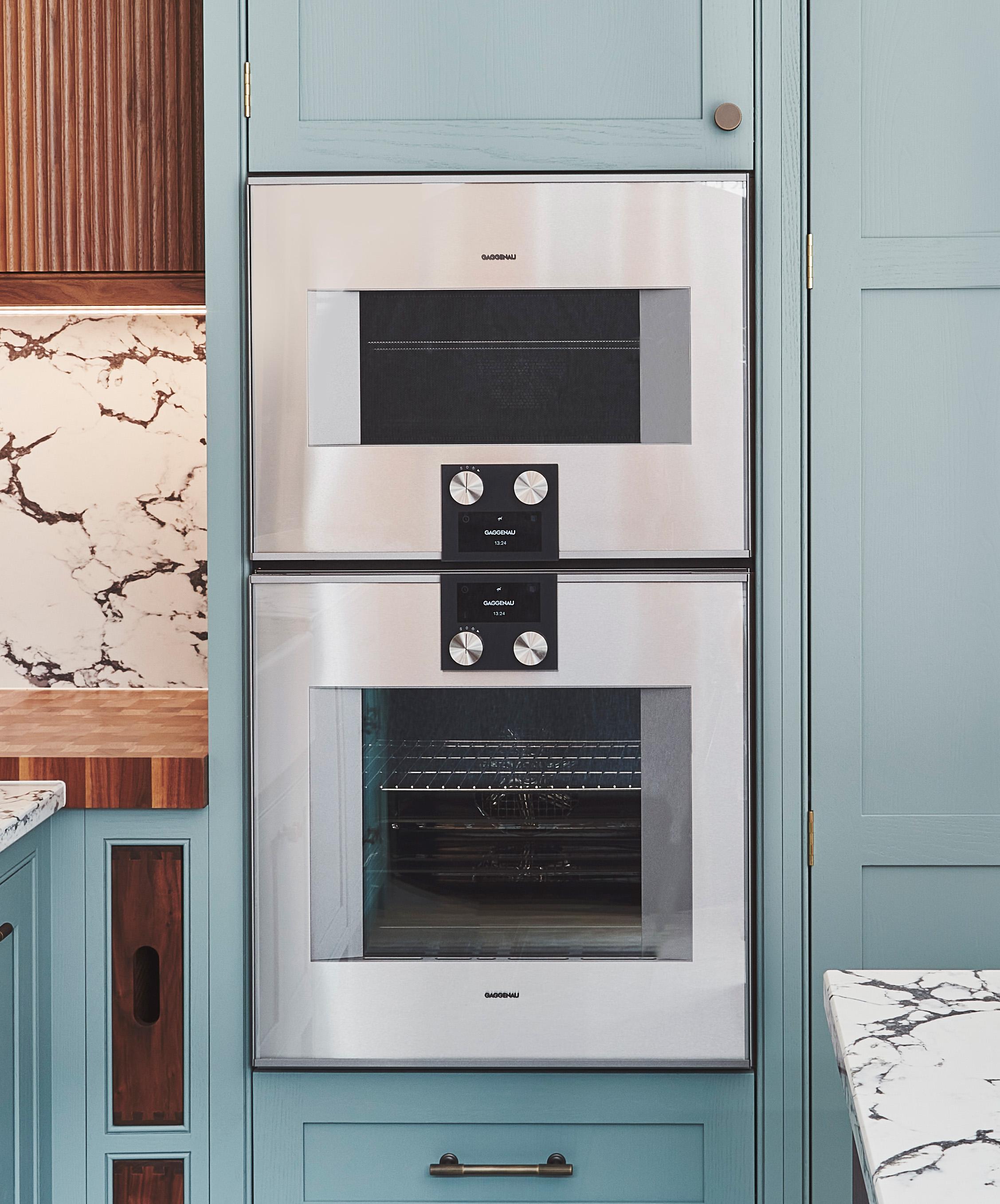 A modern kitchen with a built-in double oven set into light blue cabinetry. The oven has a stainless steel finish and digital controls. The countertop features a white and brown marble design, and there is wood paneling on the adjacent wall.