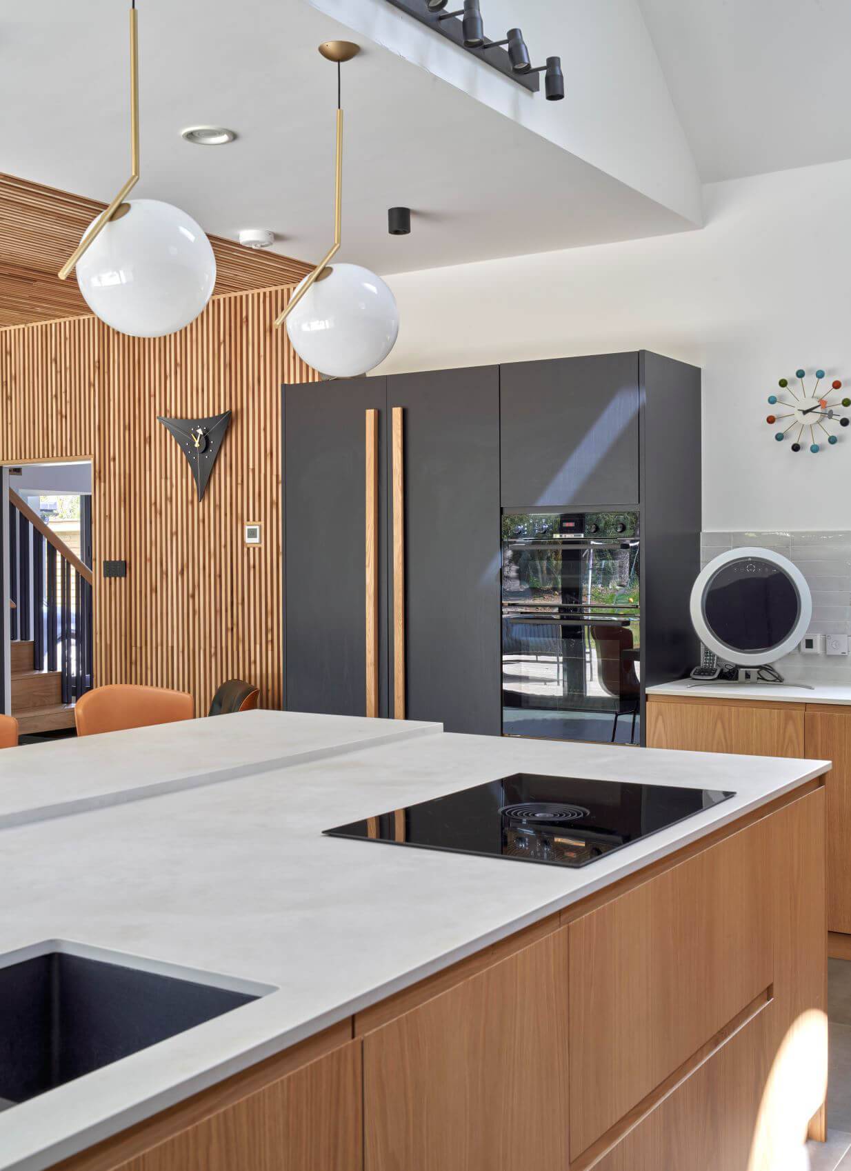 Modern kitchen with a white countertop island, built-in stovetops, and wooden cabinets. Black fridge, retro clock, and spherical pendant lights add style. Wooden wall panels and unique geometric decor accent the sleek, contemporary design