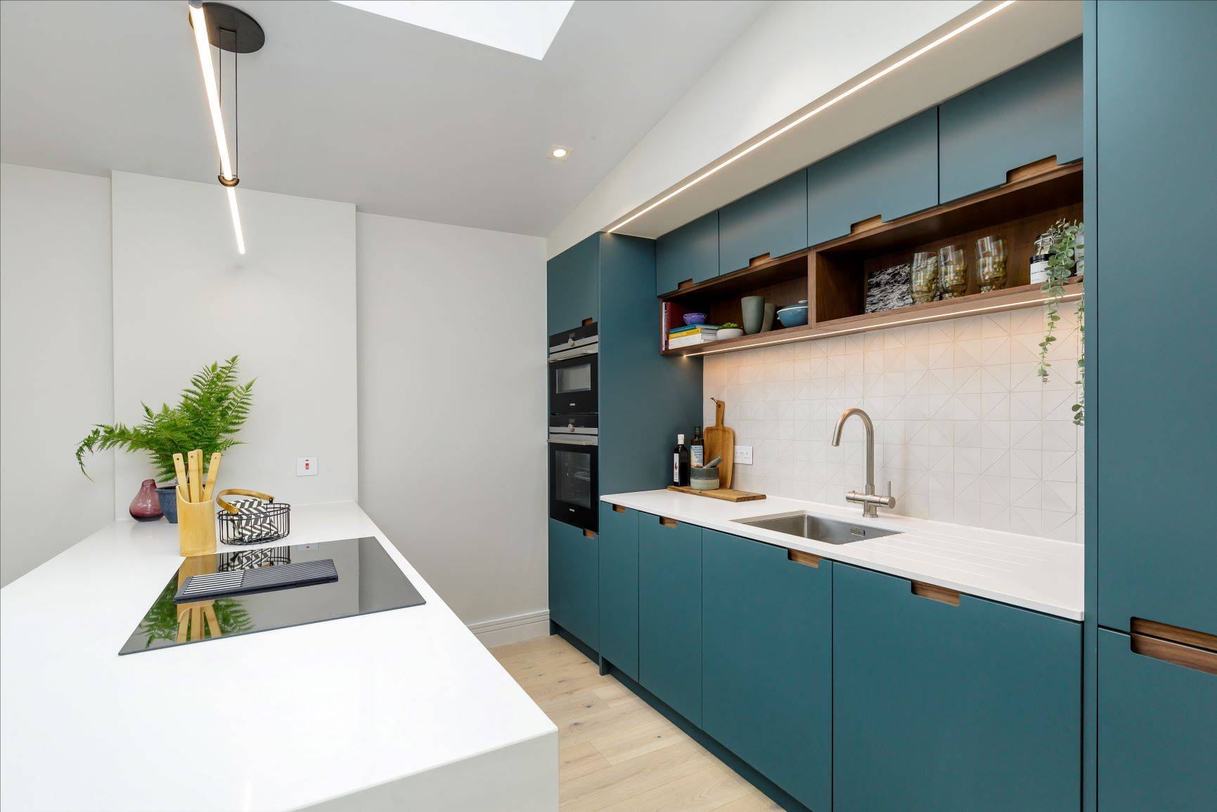 A bright kitchen with a large window, blue cabinets with glass doors, and a marble-topped island. Two wooden bar stools sit by the island, and a vase of pink roses is on the counter. A pendant light hangs above.