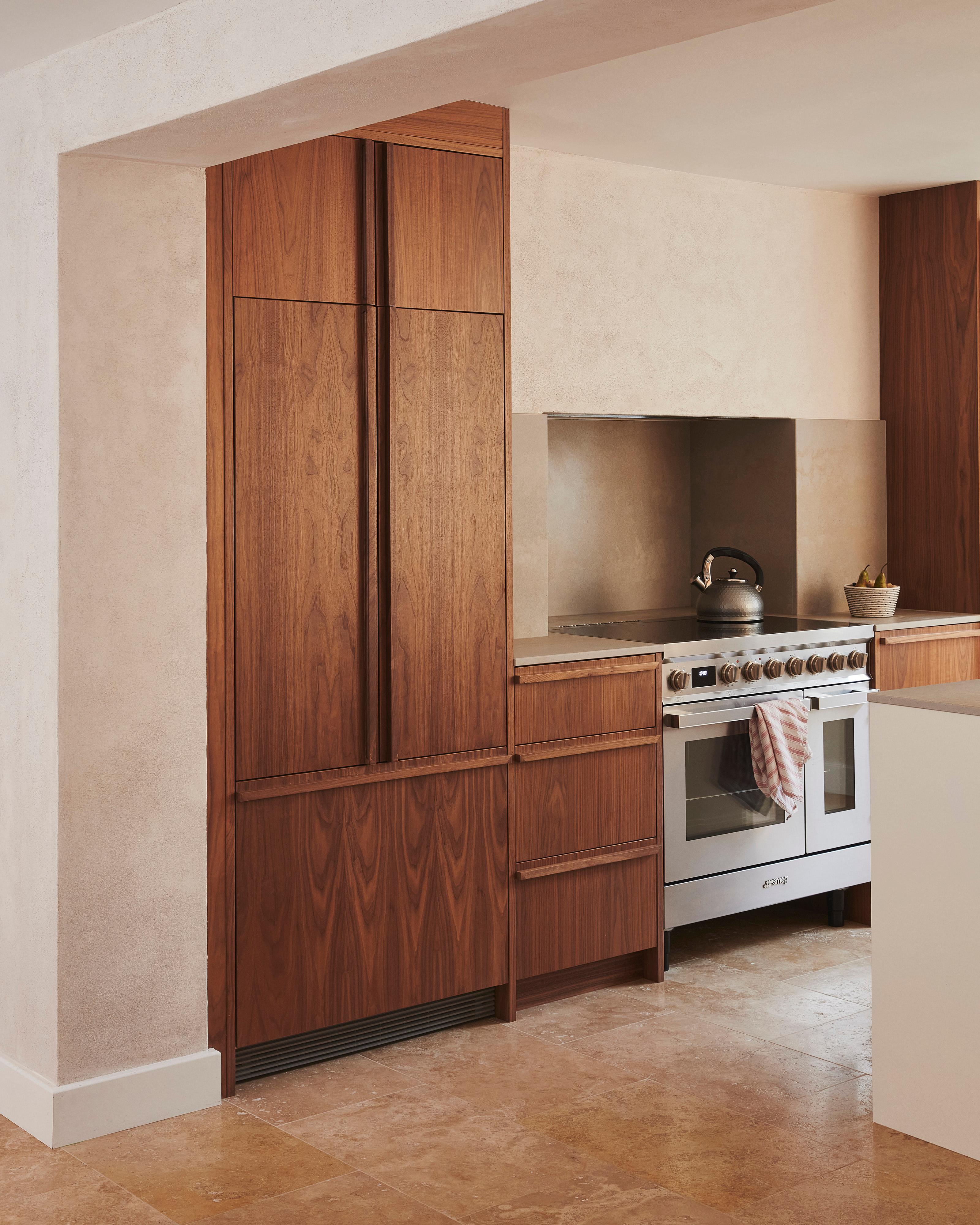 A modern kitchen with wooden cabinets, a stainless steel oven, and a white countertop. A kettle sits on the stovetop, and a small plant is placed on the counter next to the oven. The floor is made of light beige tiles.