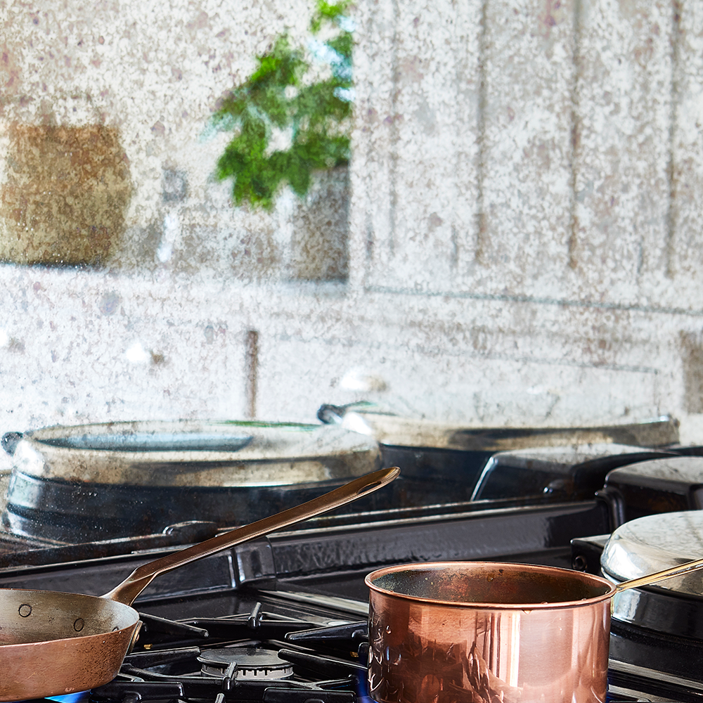 A close-up of a stovetop with pots and pans, including a copper pot, in a kitchen. The background is slightly blurred, showing white cabinets and a plant reflected on a greasy backsplash.